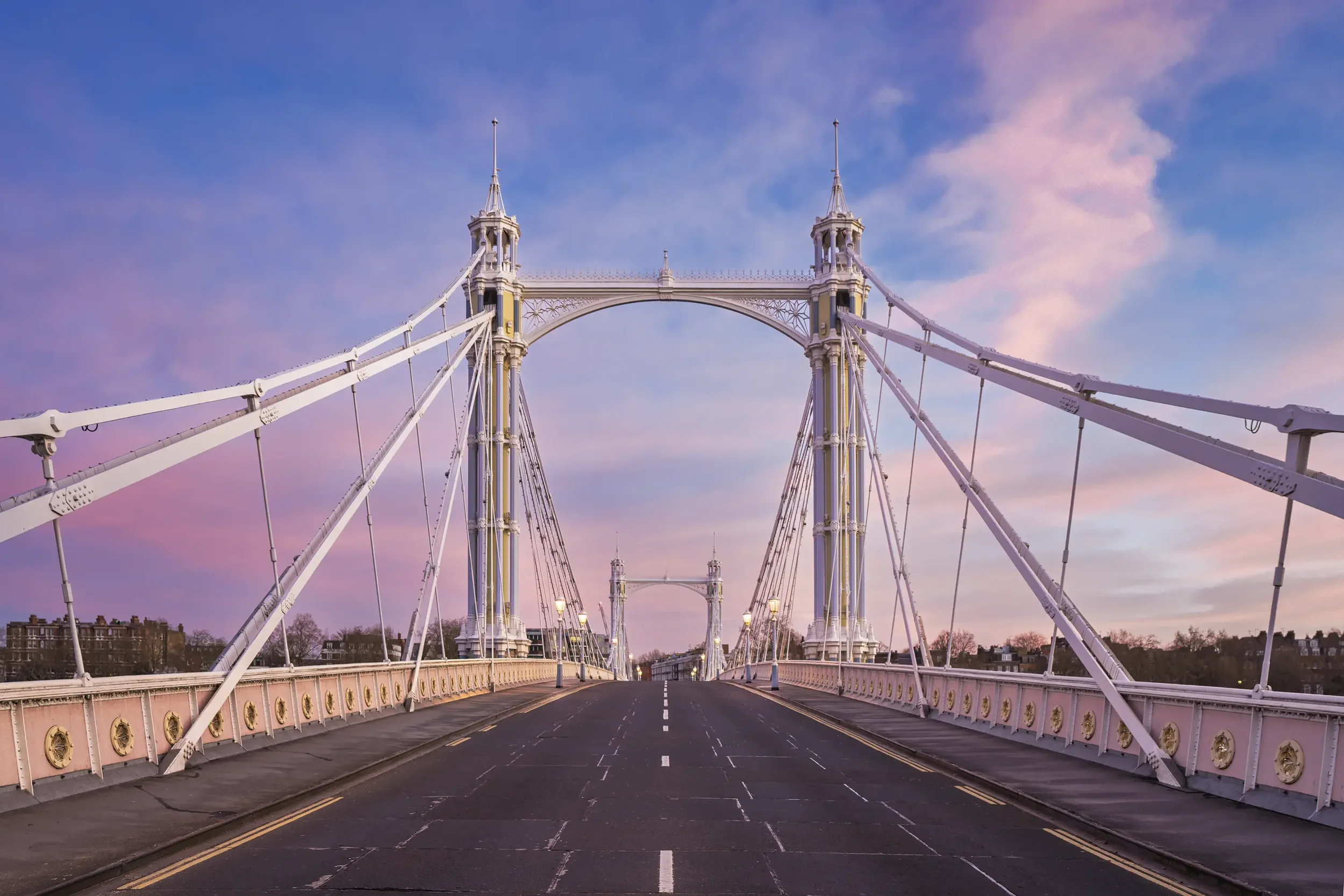 A white suspension bridge with tall towers and decorative railings, empty roadway, against a colorful sky with pink and blue hues, and cityscape in the background.