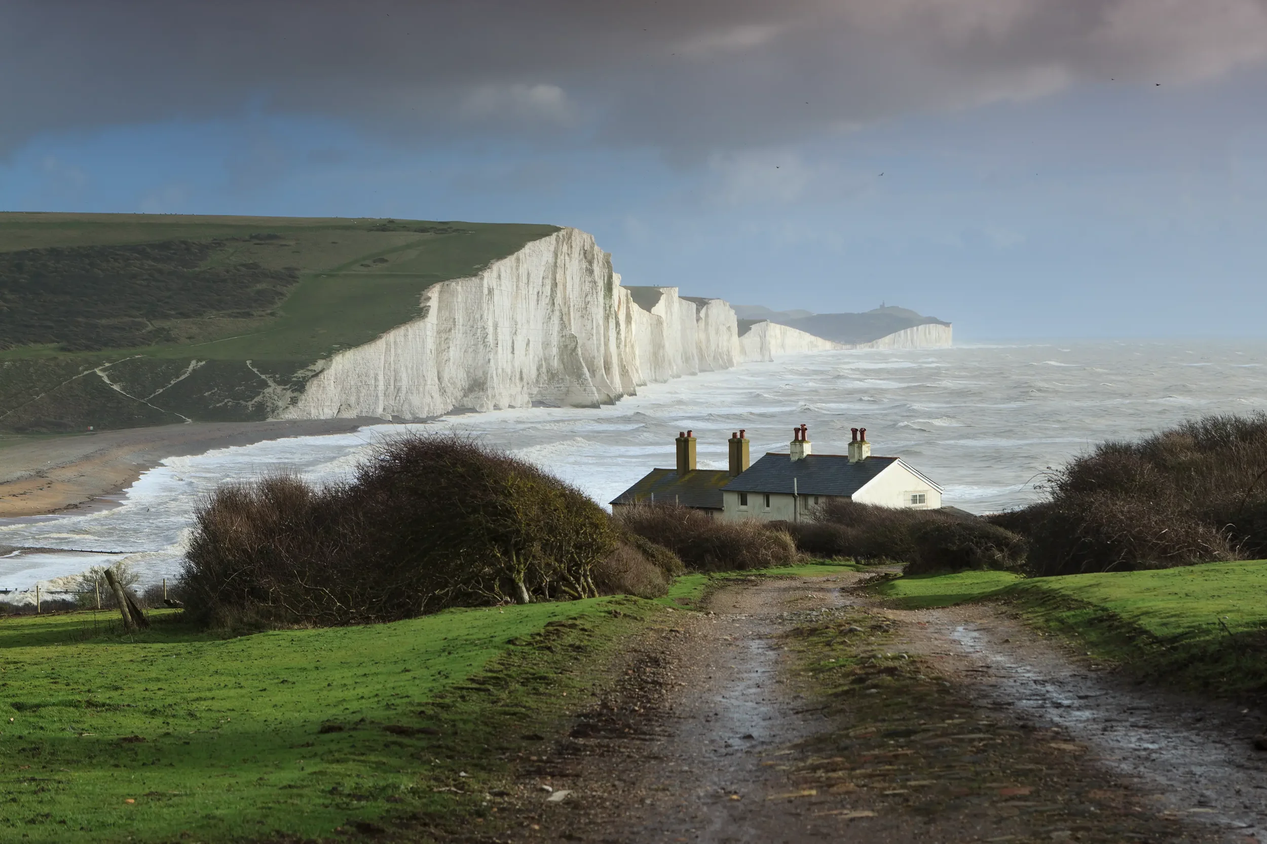 White chalk cliffs along the coast with grassy tops and a small beach area underneath. Several houses with chimneys are in the foreground on a muddy path leading towards the cliffs. The sky is overcast with dark clouds.