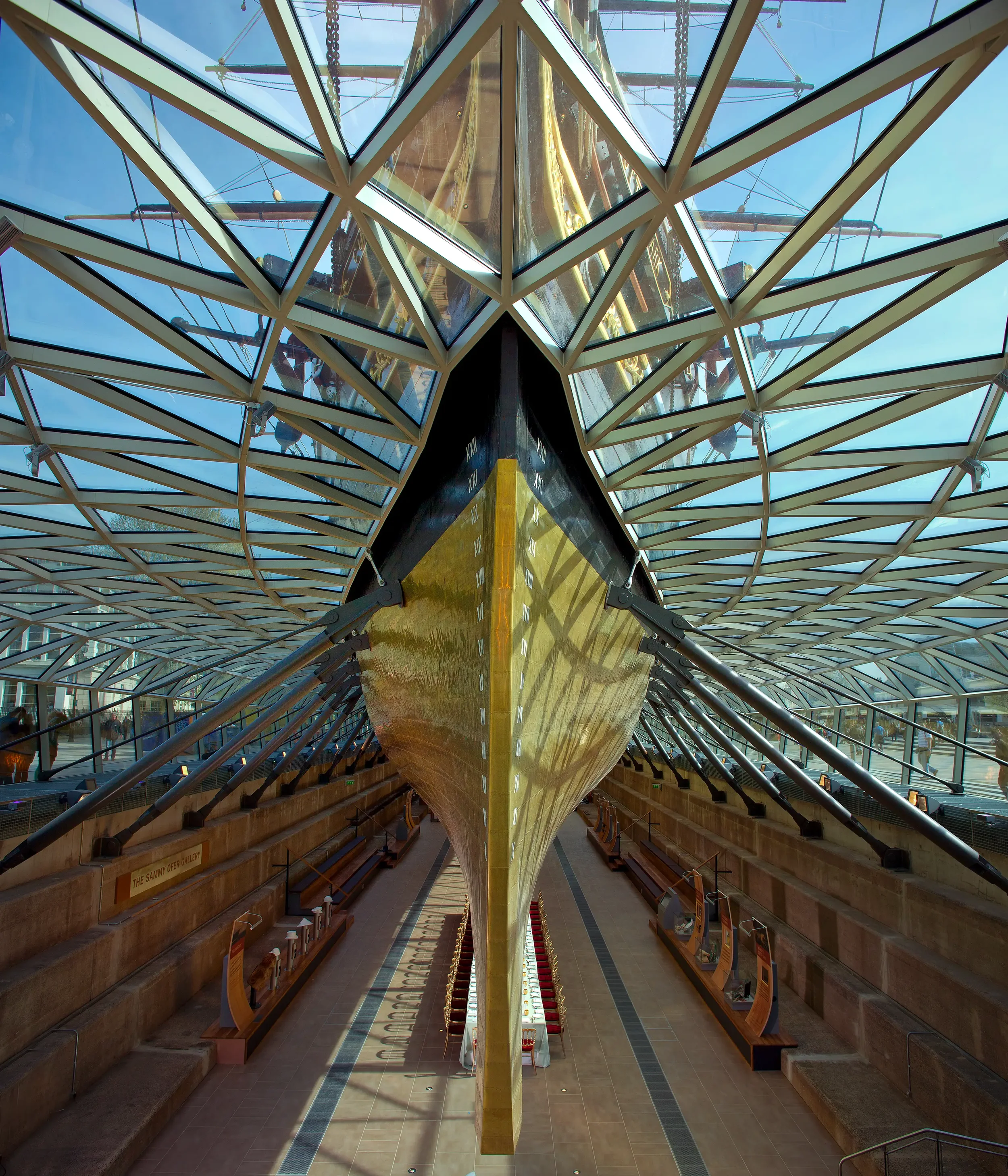 The image depicts the submerged bow of the RMS Titanic exhibit in the Titanic Belfast Museum, viewed from below through a glass ceiling with a metal framework, with a clear blue sky visible outside.