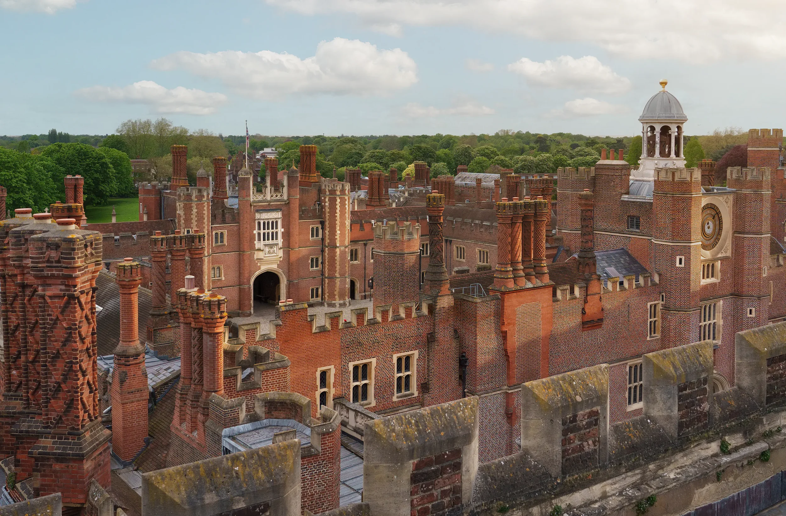 A historic brick castle with numerous chimneys and a large clock tower surrounded by green trees under a cloudy sky.