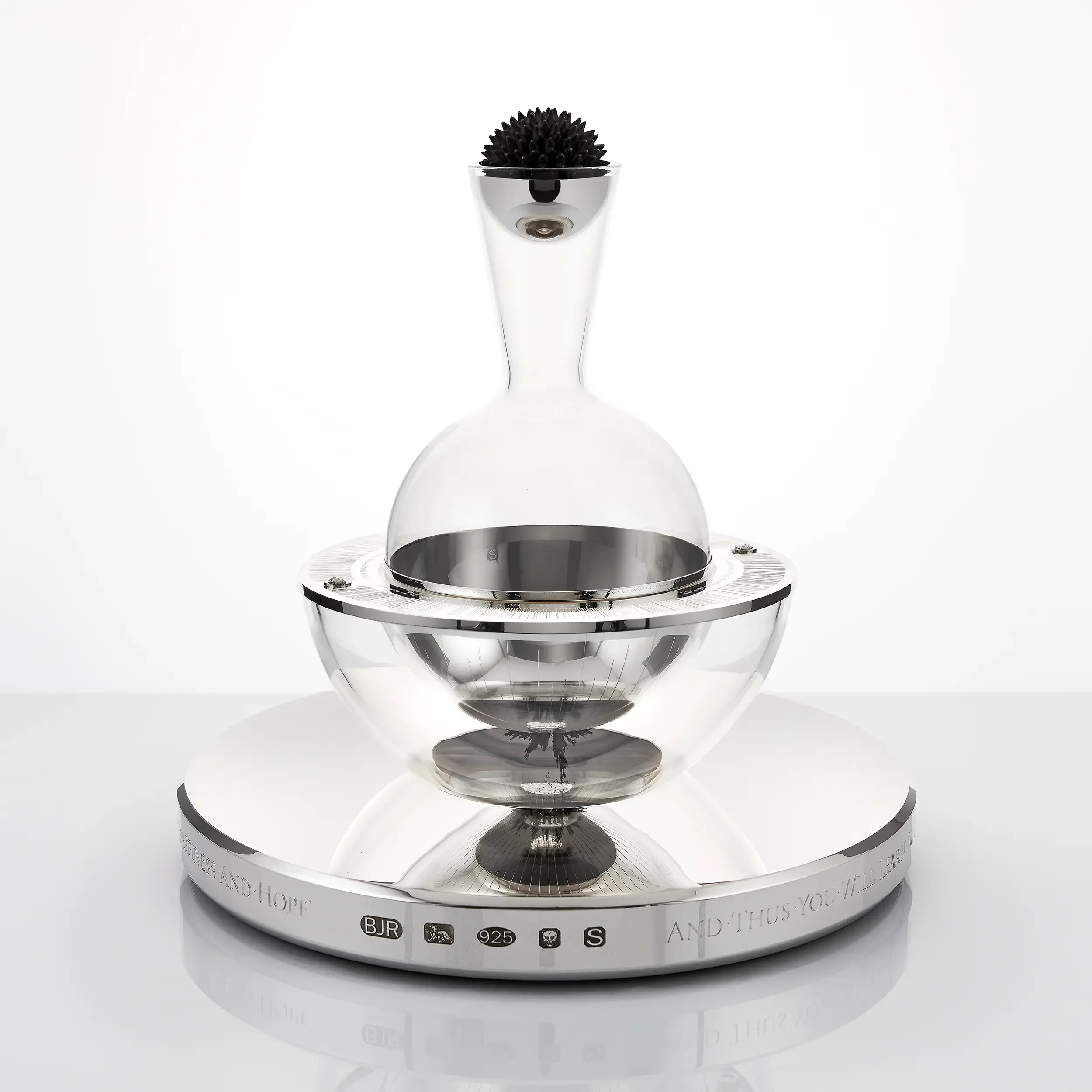 A clear glass award with a black spiky object inside, mounted on a round white base with engraved text and various symbols around the edge.