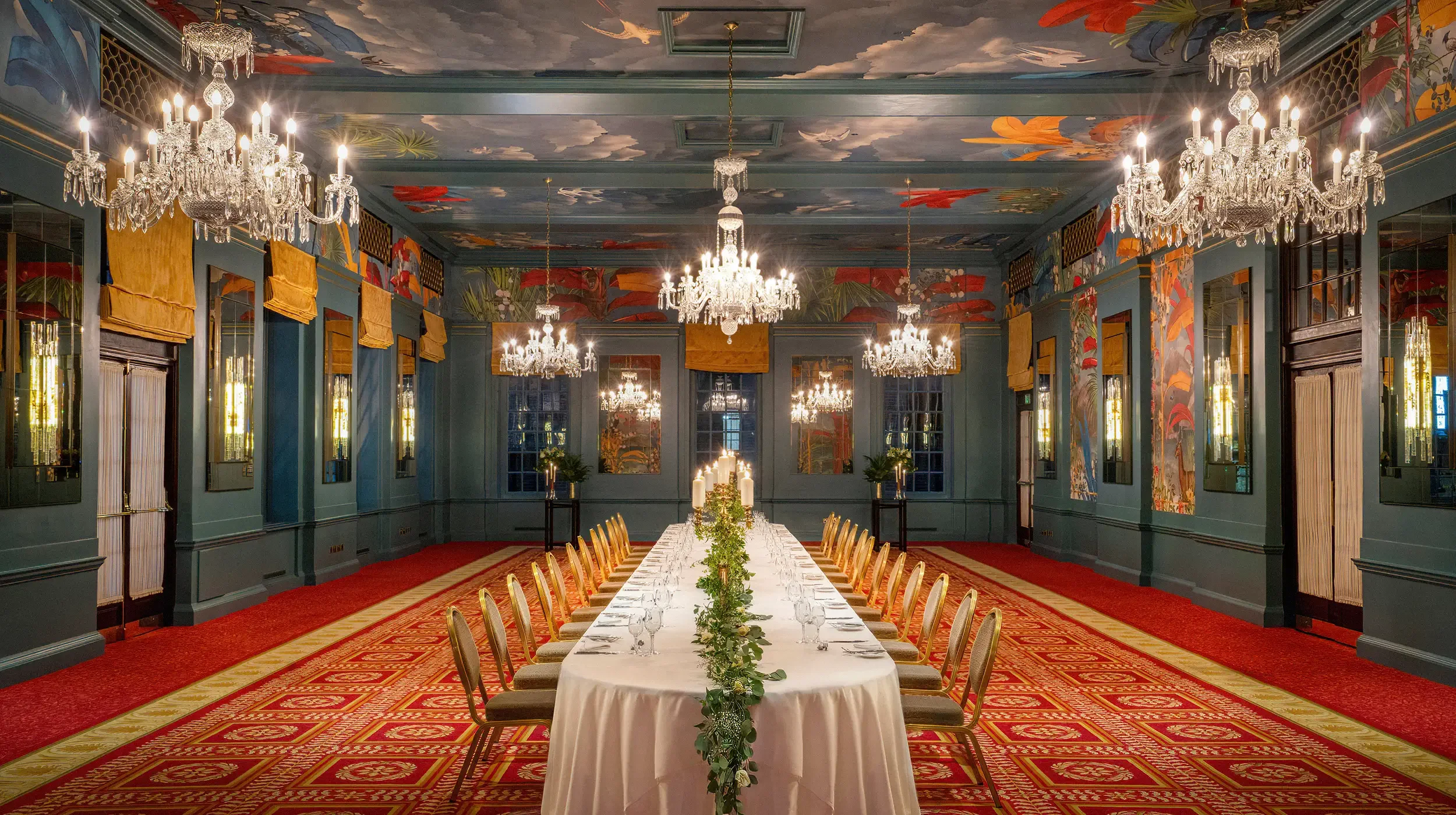 Elegant dining room with long table set with glassware and a greenery centerpiece, surrounded by gold chairs, ornate chandeliers, and decorated walls with floral murals and gold accents.