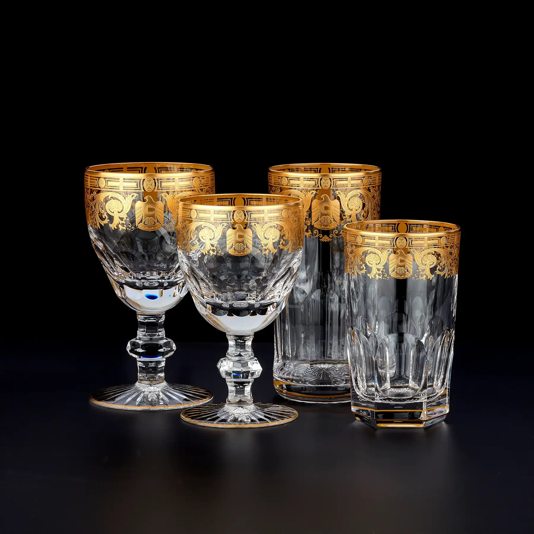 Set of four intricately designed crystal and gold-rimmed drinking glasses displayed against a black background, with two larger glasses and two smaller ones.