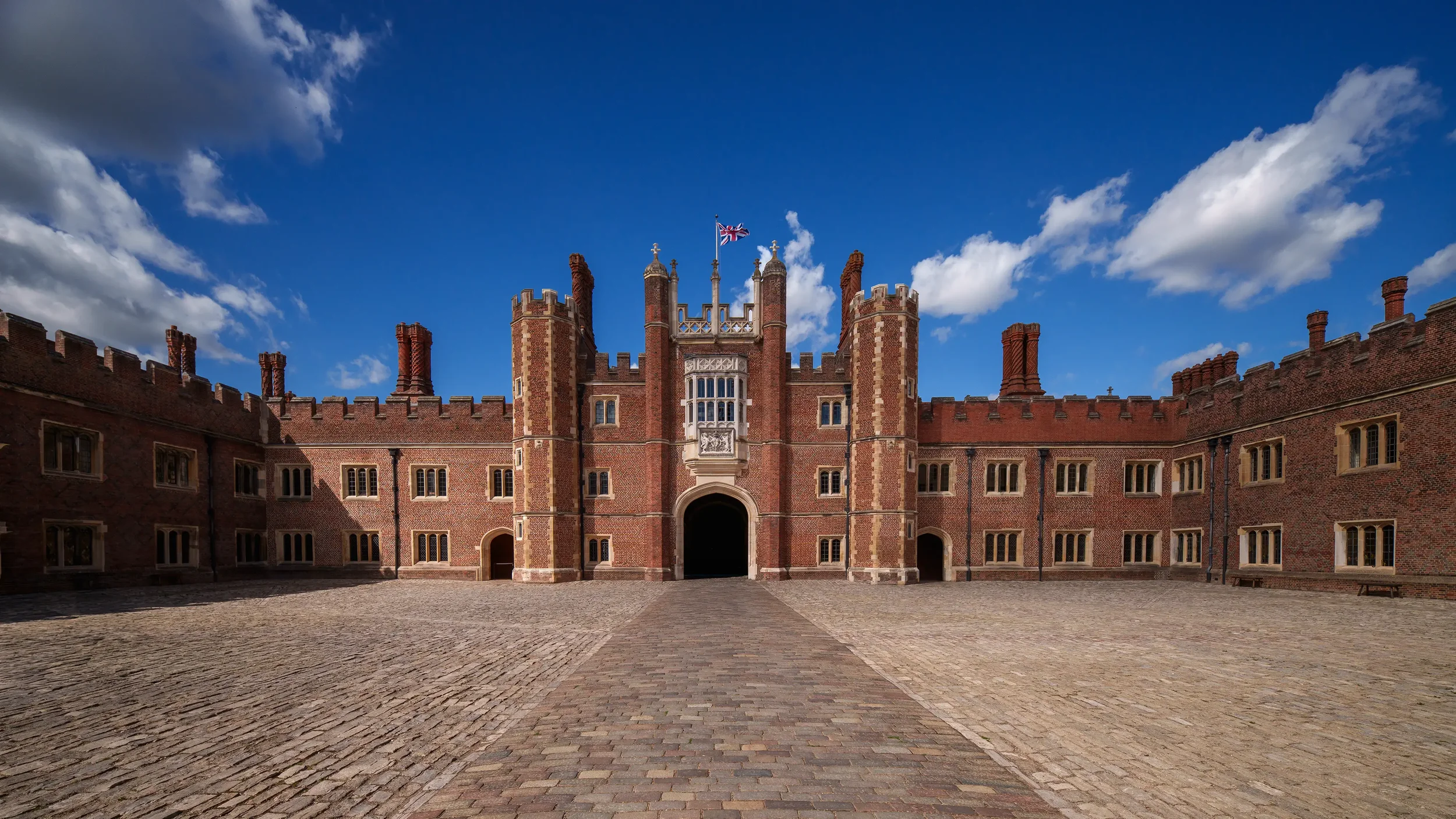 A large, historic brick castle with multiple towers, battlements, and a central entrance, flying a Union Jack flag under a partly cloudy blue sky.