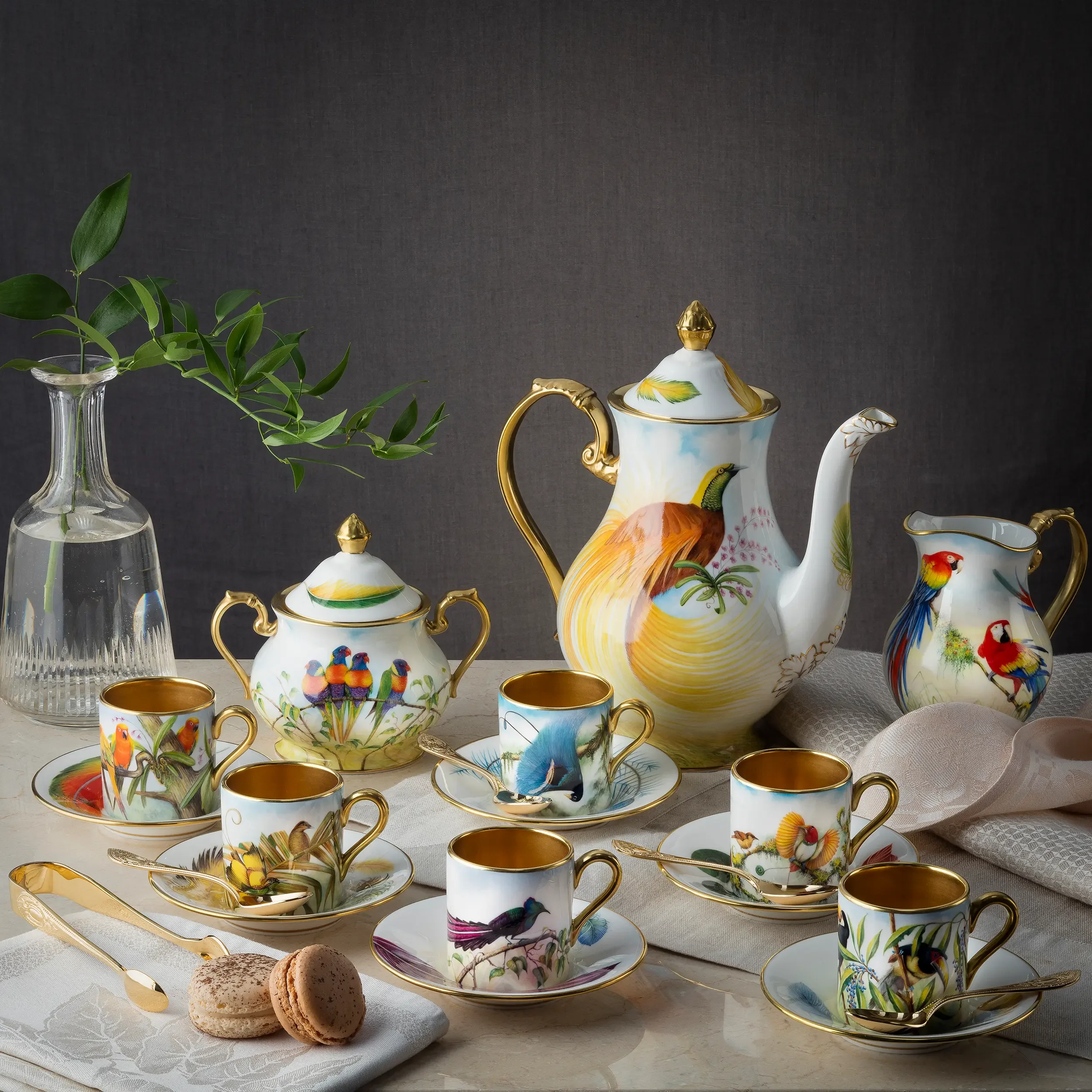 A decorative tea set with painted bird and floral designs, including a teapot, cups, saucers, and sugar bowl with golden accents, displayed alongside a glass vase with green leaves, on a white tablecloth against a dark background.