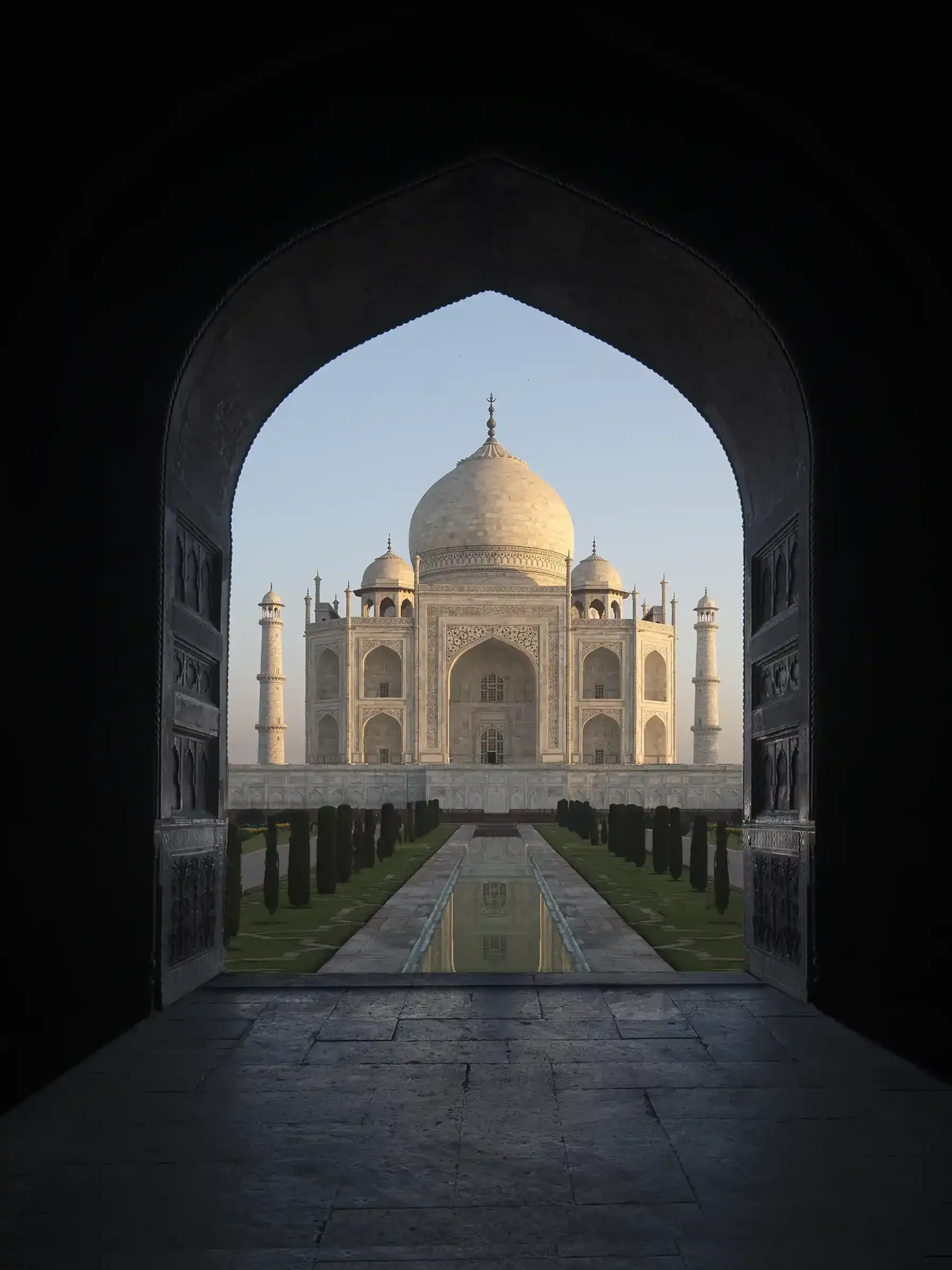 View of the Taj Mahal through a dark ornate archway, with a reflection pool and pathway leading towards the monument.