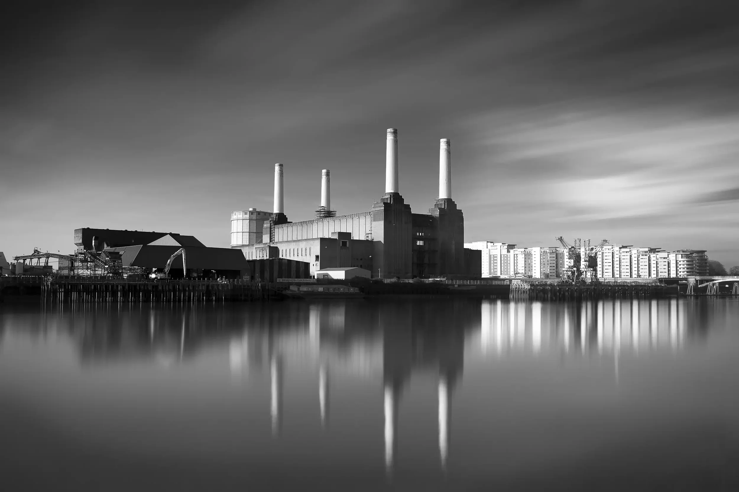 Black and white photo of a power plant with three smokestacks near a body of water, reflecting the building and sky.