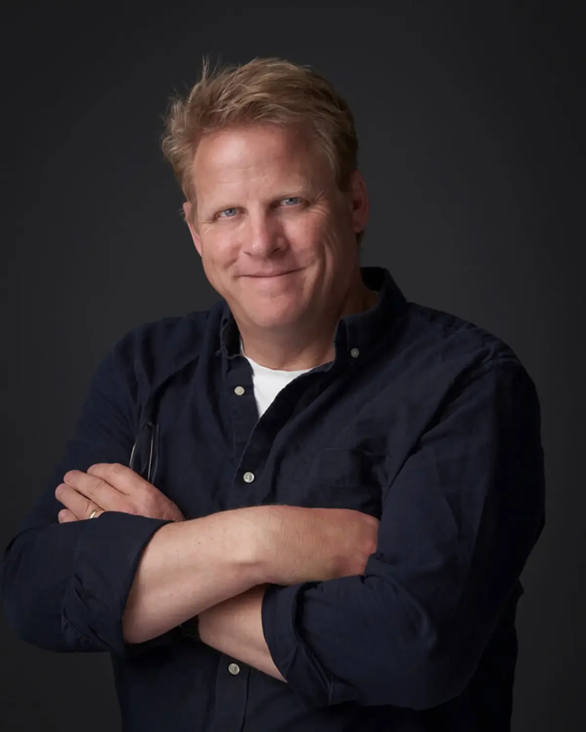 A middle-aged man with light brown hair and blue eyes, smiling, wearing a black button-up shirt with sleeves rolled up, crossed arms, standing against a dark background.