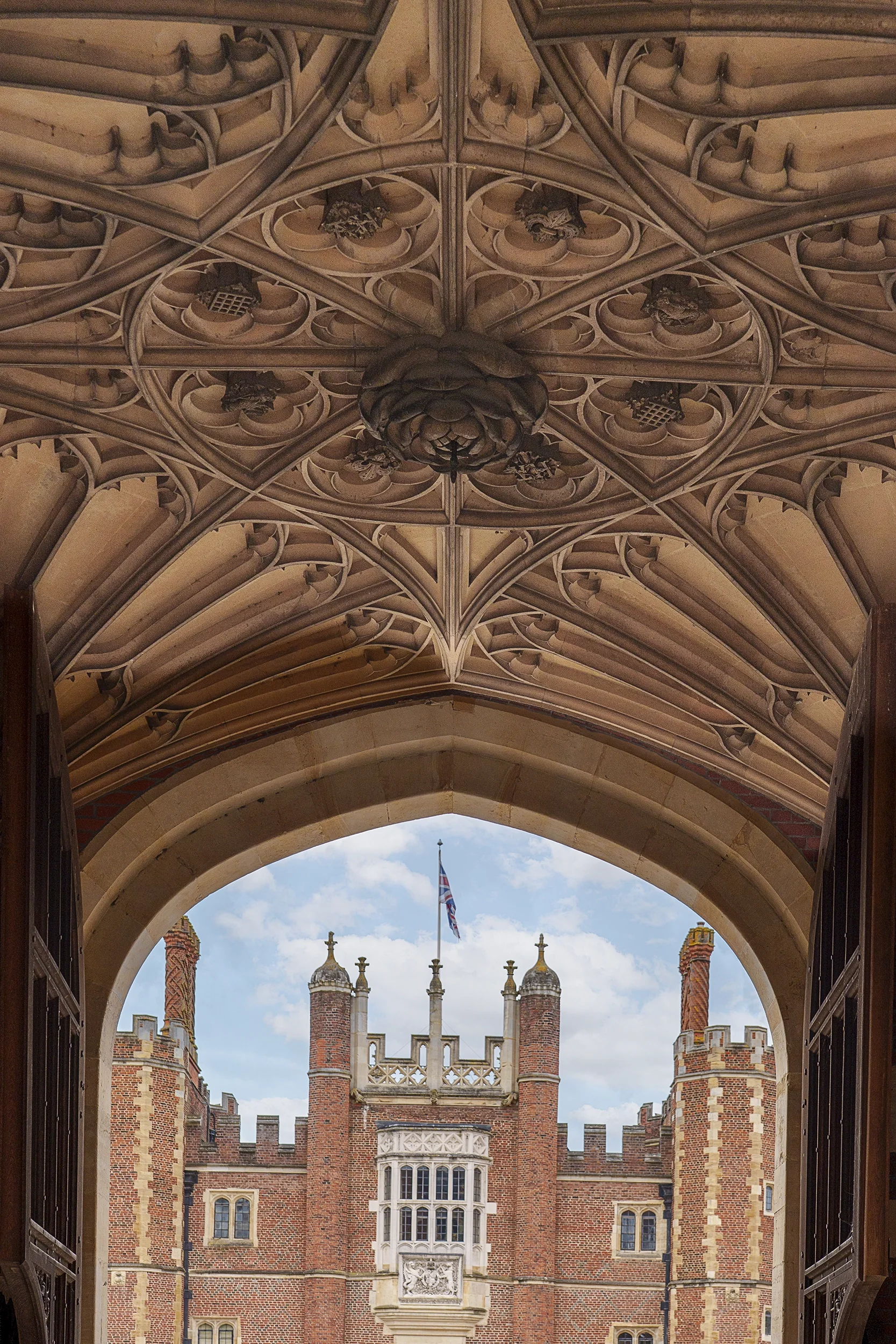 View from a Gothic-style archway looking out towards a historic red brick castle with turrets, chimney stacks, and a flag flying on top, under a cloudy sky.