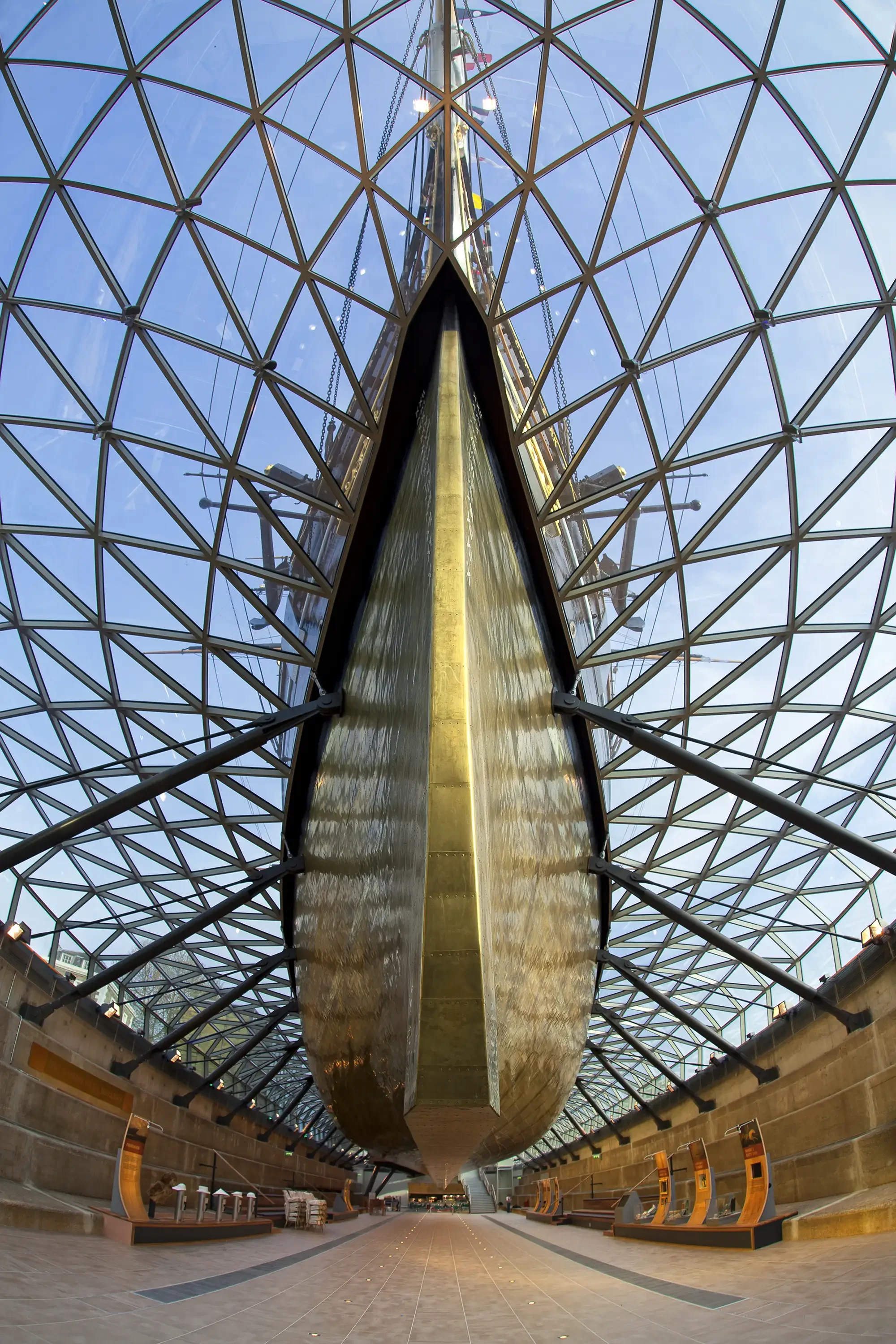 Interior of a modern structure with a large, boat-shaped, gold-colored vessel suspended from a glass and metal dome ceiling, with a tiled floor and seating areas along the sides.