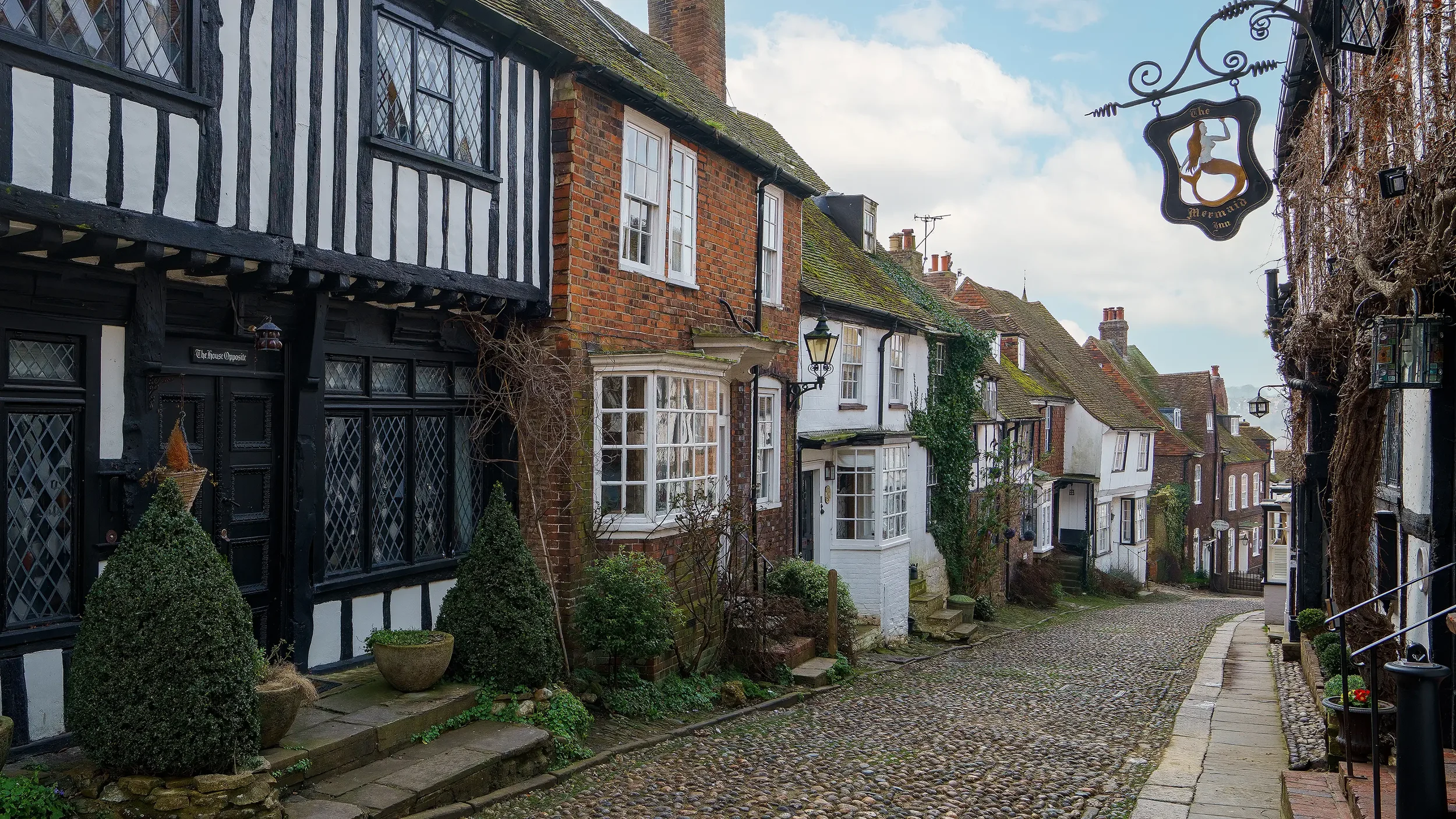 A scenic street view of old European-style houses with cobblestone pavement and traditional architecture, including timber-framed and brick facades, with cloudy sky overhead.