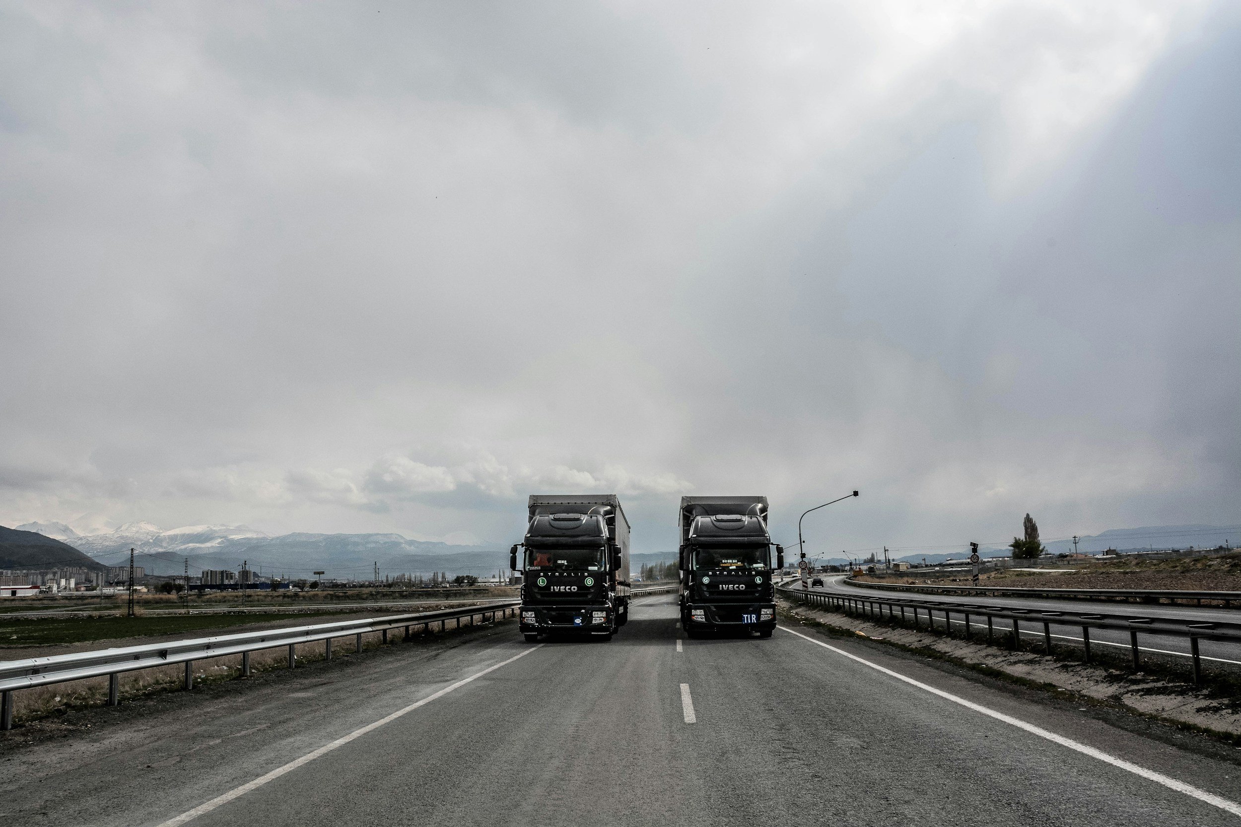 Two large black trucks on a highway, with mountains in the background and a cloudy sky overhead.