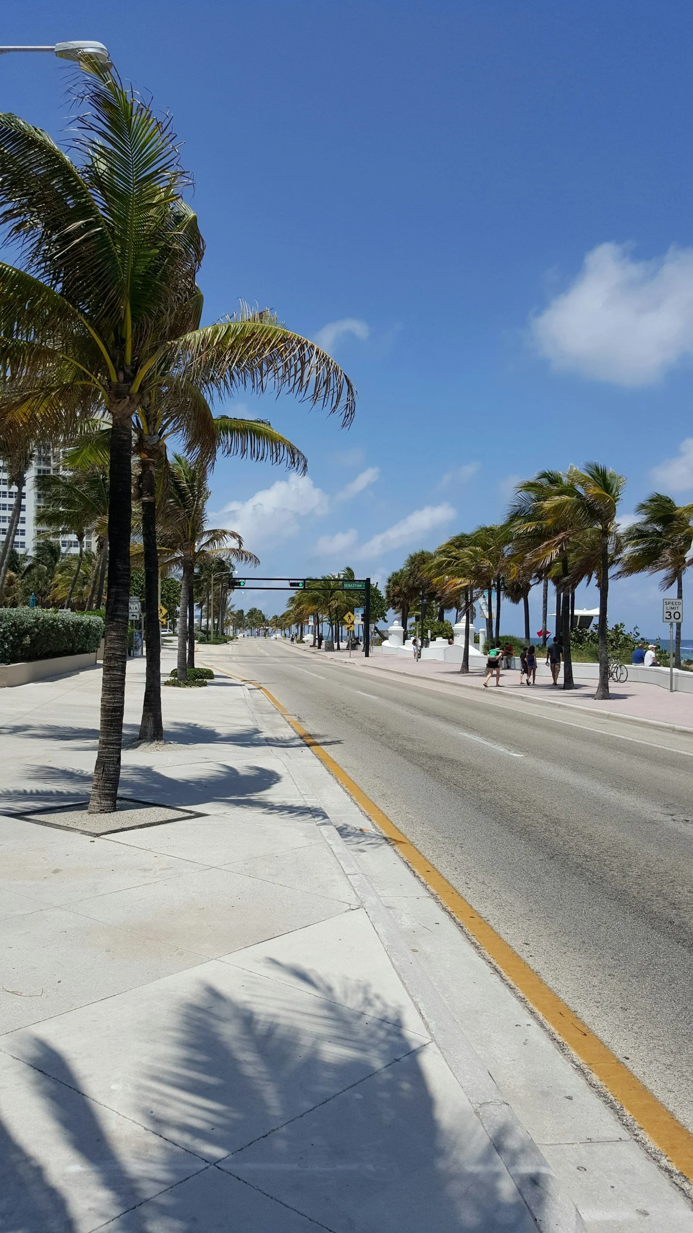 A sunny street lined with palm trees along a sidewalk, with a few people walking and a clear blue sky overhead.