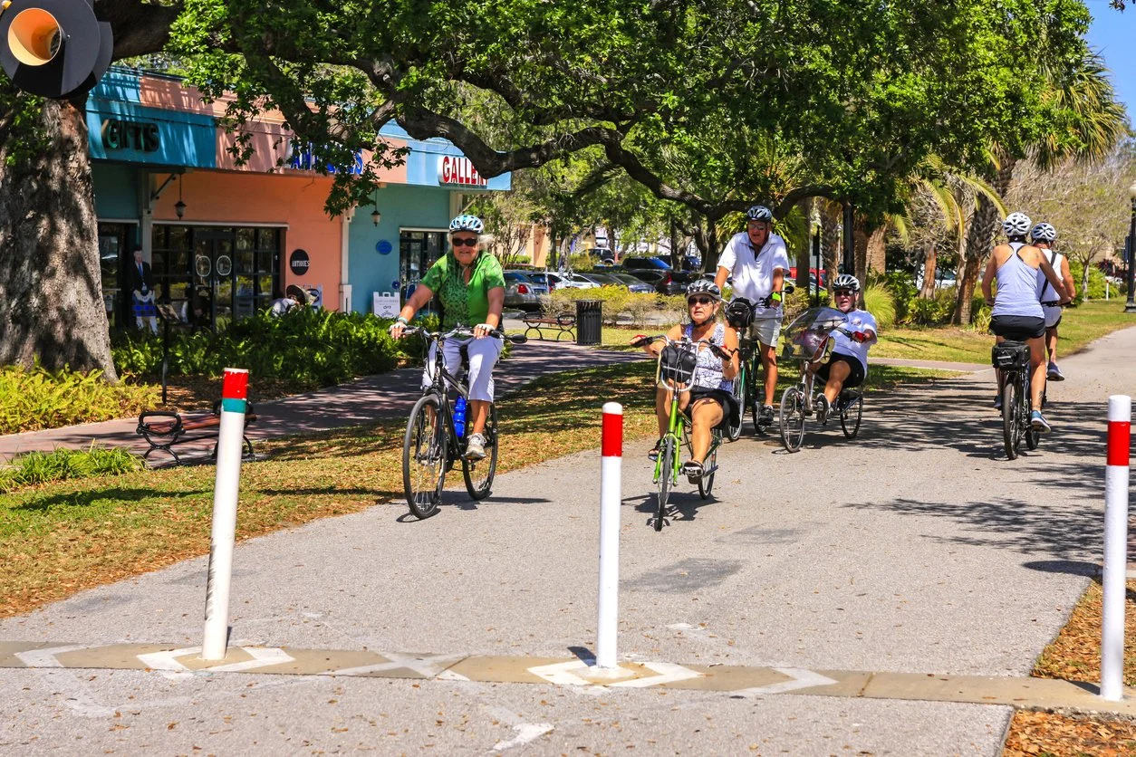 People riding bicycles on a park pathway under a large tree with green foliage, with a pastel-colored building in the background.