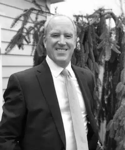 A smiling man in a suit standing outdoors with a wooden house and trees in the background.