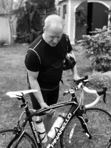 A person wearing a black shirt stands outdoors in a backyard, holding a bicycle.