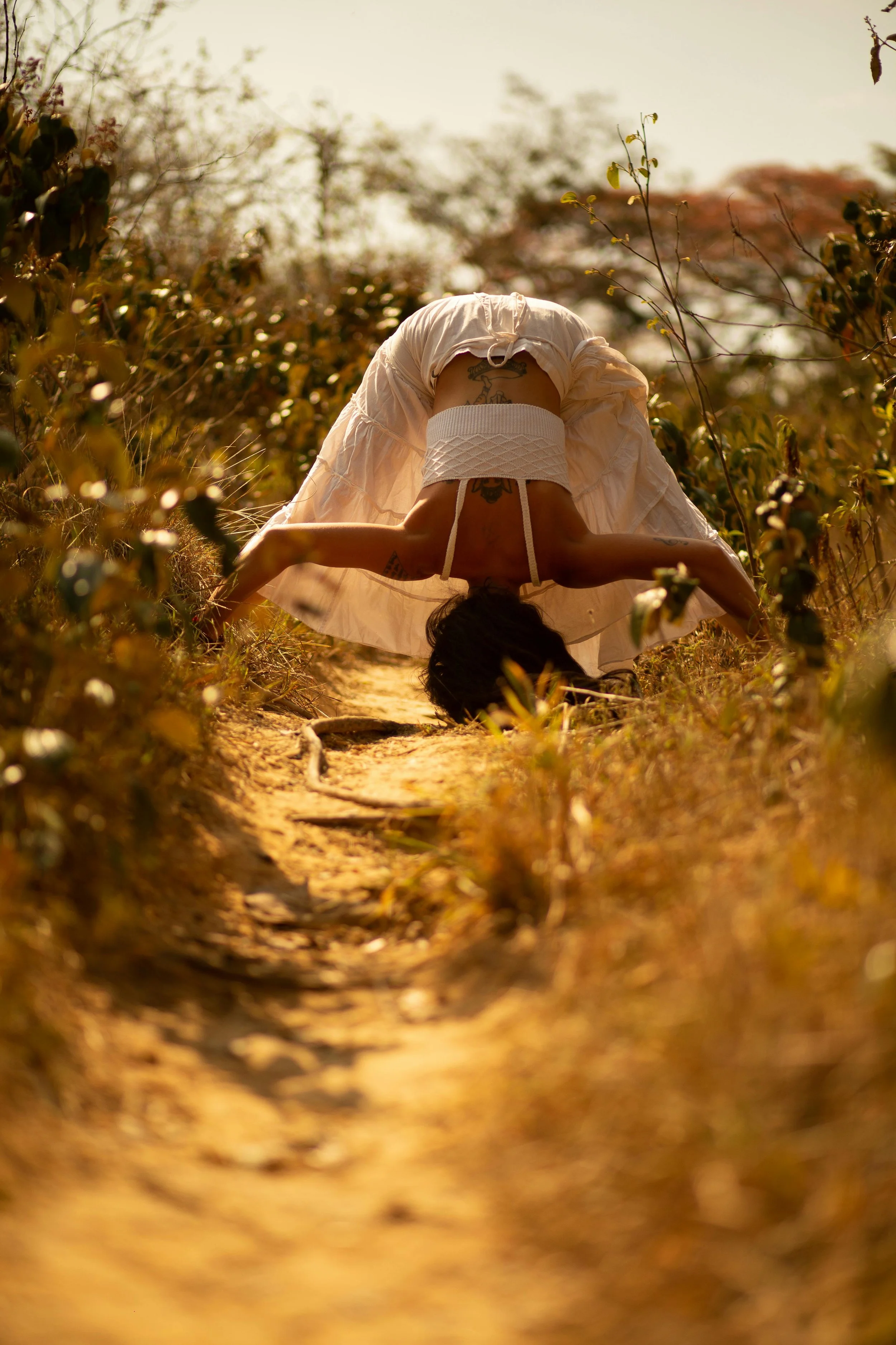 Person in white dress performing a headstand or yoga pose outdoors on a dirt path surrounded by plants and trees during golden hour.