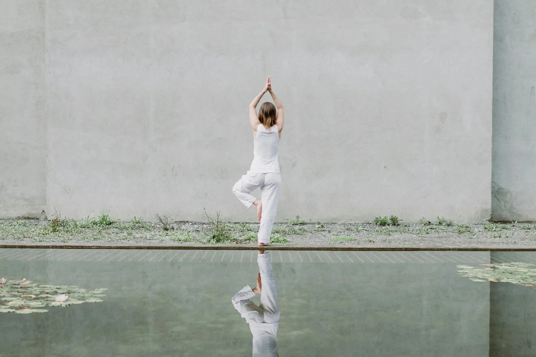 A woman practicing yoga outdoors in a yoga pose called tree pose, standing on one leg with hands pressed together overhead, reflected in a still pond with water lilies, against a plain concrete wall.