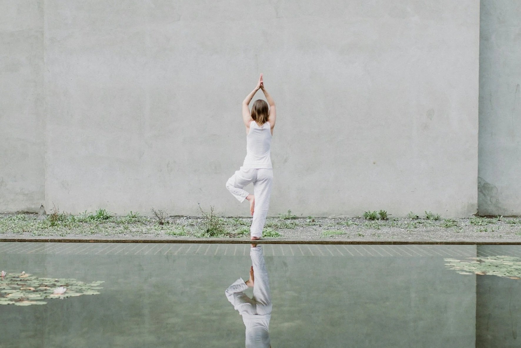 A woman practicing yoga outdoors, standing on one leg in a tree pose with her hands in prayer position above her head, reflected in a body of water.