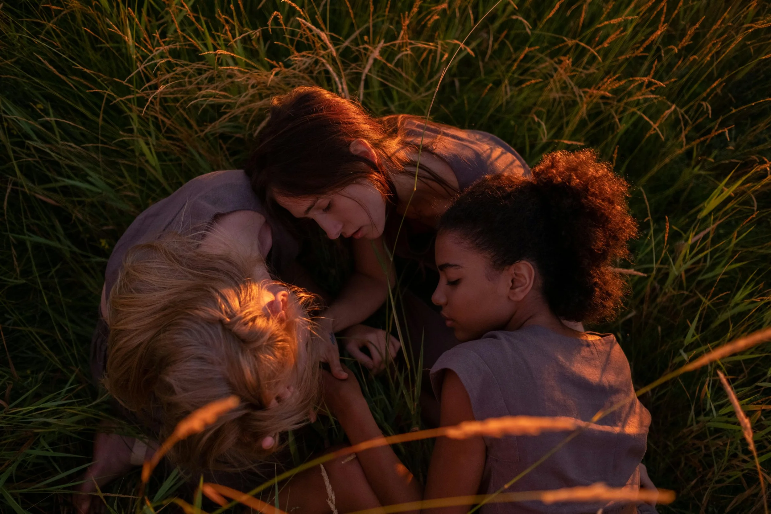 Adult woman and four children lying close together in tall grass during sunset, with their eyes closed and heads touching.