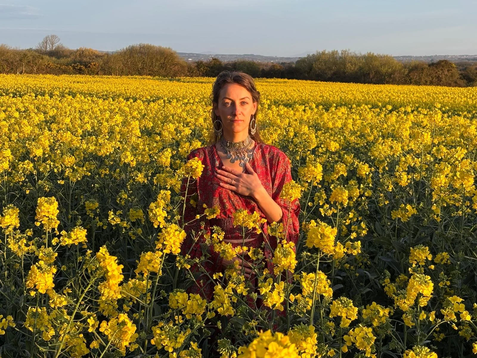 A woman in a red dress with jewelry, standing in a field of yellow flowers during the daytime.