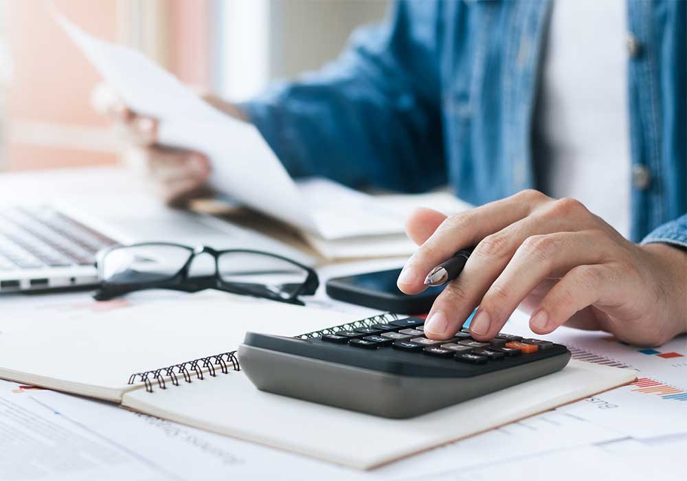 Person using a calculator at a desk with papers, a notebook, glasses, a smartphone, and a laptop.