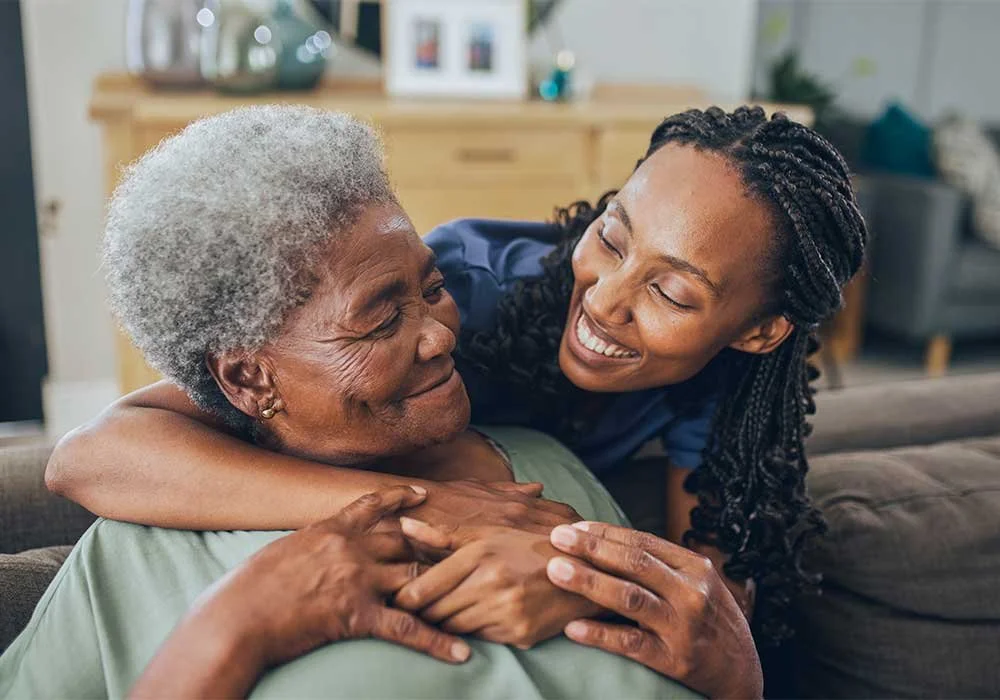 Young woman hugging elderly woman and smiling warmly, inside a cozy living room.