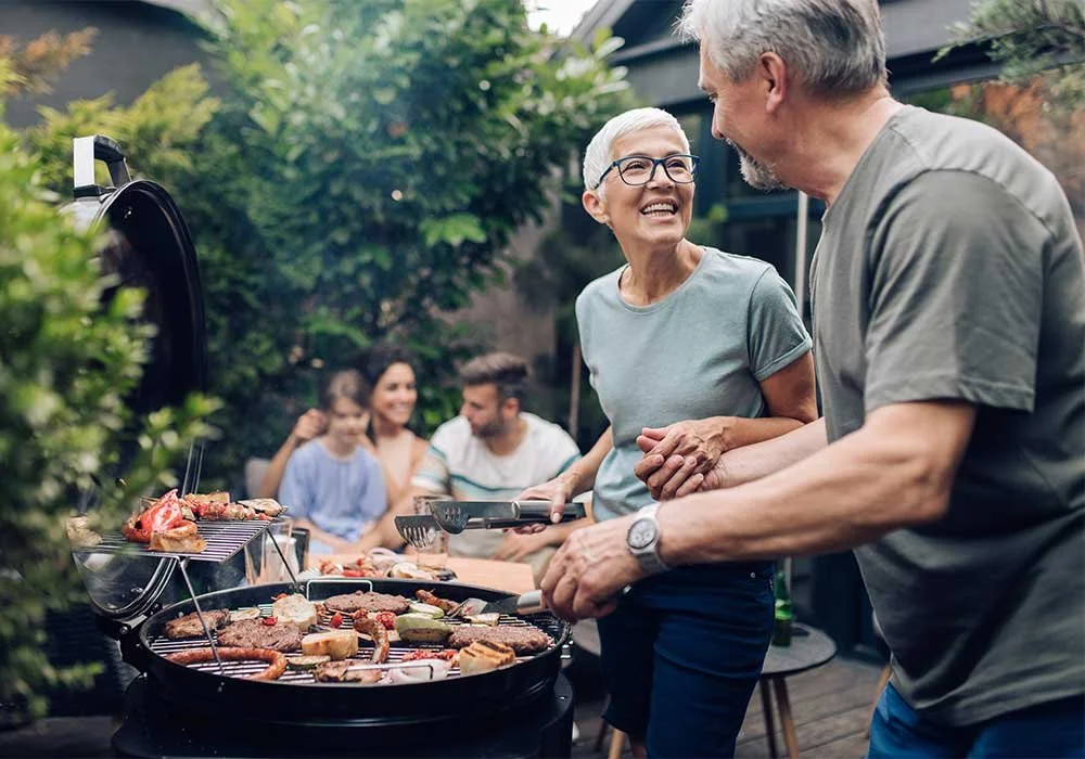 An elderly couple laughing and talking while cooking on a backyard grill during a gathering, with a group of people sitting and talking in the background.