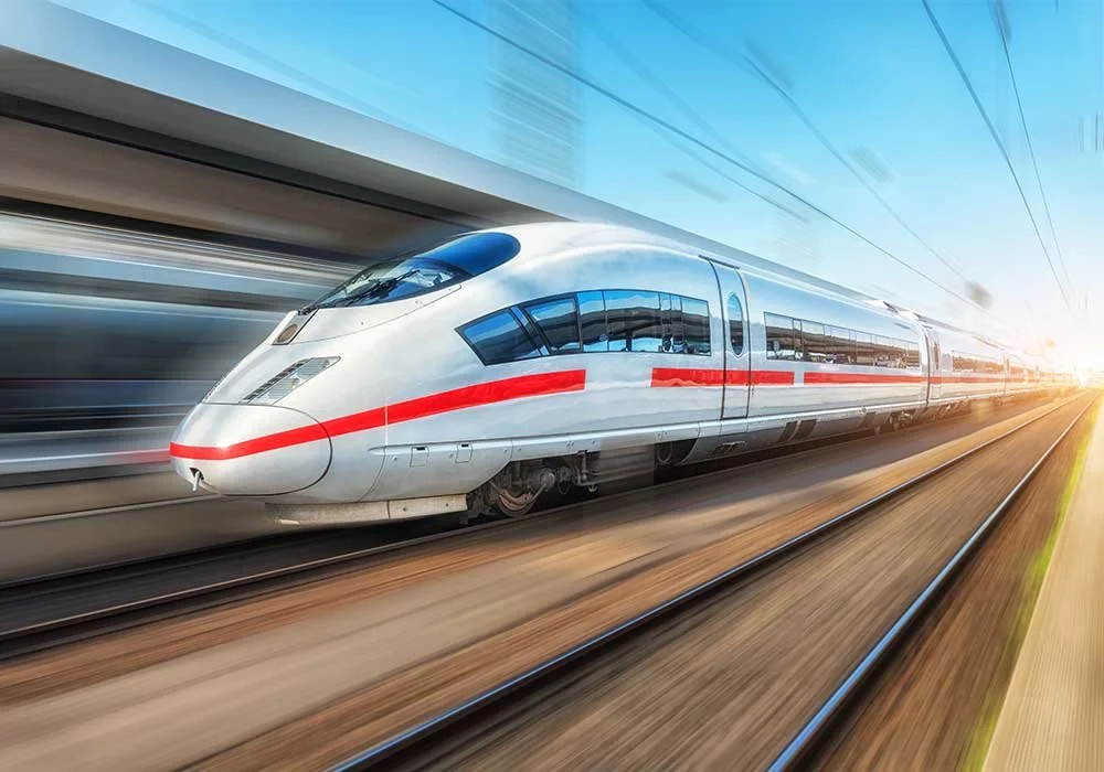 A modern, white bullet train with red stripes traveling on railway tracks, blurred background, and sunlight at the horizon.
