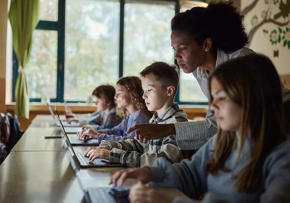 A teacher assists students working on laptops in a classroom with large windows and green curtains.