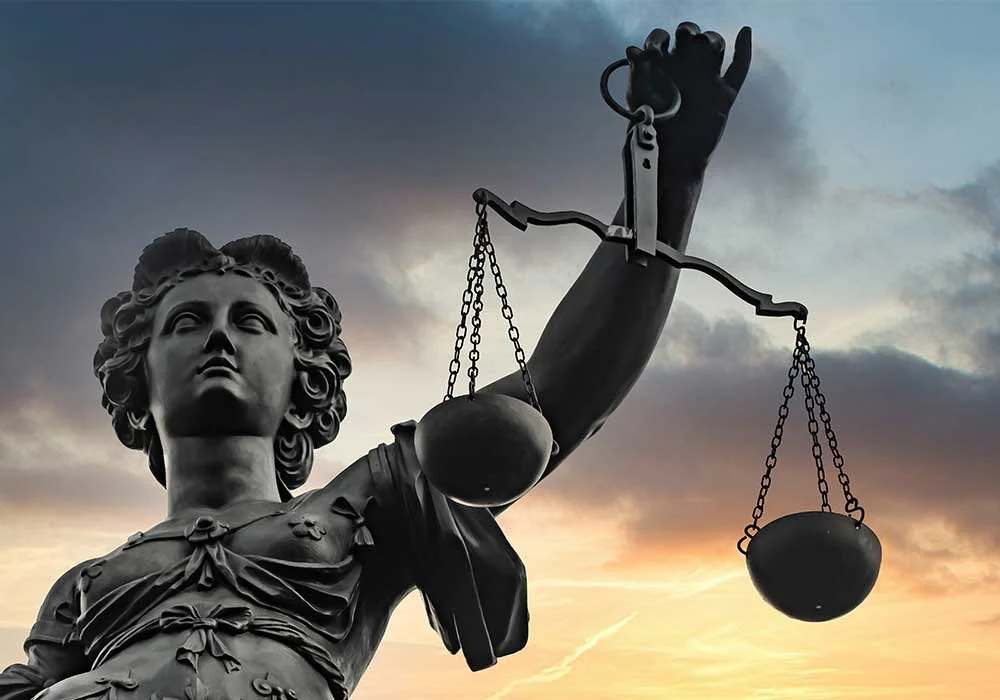 Dark statue of Lady Justice holding a set of scales against a sunset sky with clouds.