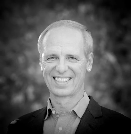 Black and white portrait of Dirk Langer smiling with light hair, wearing a suit jacket and shirt.