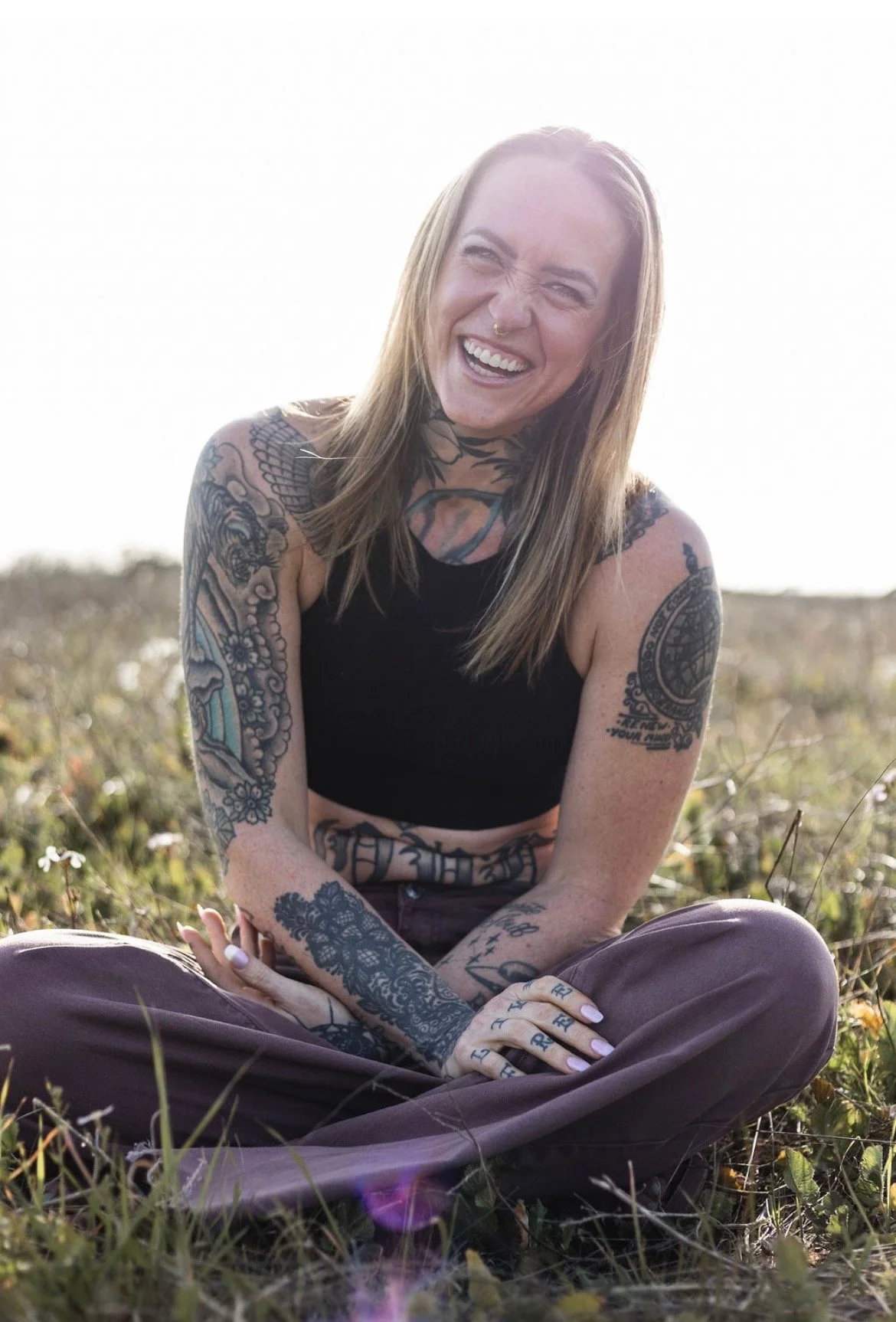 A woman with tattoos on her arms, shoulders, and neck, wearing a black sleeveless top and brown pants, sitting outdoors in a field with grass and wildflowers, laughing with her eyes closed.