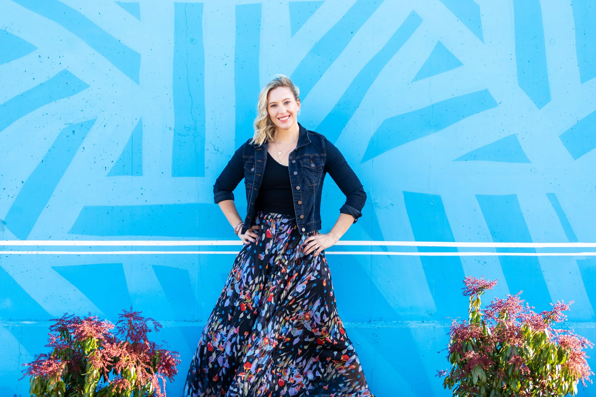 Mary Allison Jensen, LICSW, standing confidently in a floral maxi skirt and denim jacket against a bright blue mural — founder of A Legacy of Healing and host of The Change That Changes Everything free podcast series.
