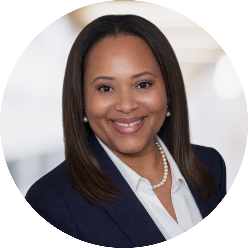 Professional portrait of a woman with shoulder-length dark hair, wearing a navy blazer, white blouse, pearl necklace, and pearl earrings, smiling in an office setting.