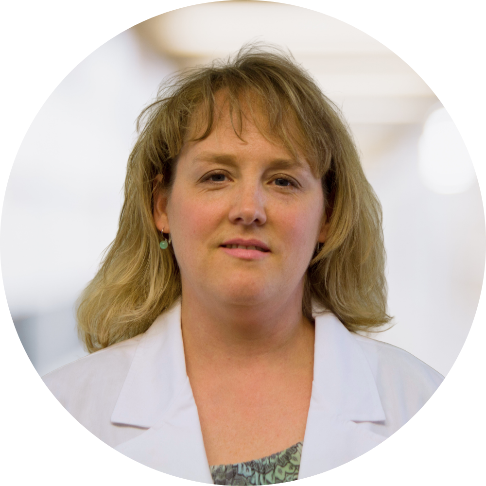 A woman with shoulder-length blonde hair, wearing a white coat, standing indoors with a blurred background.
