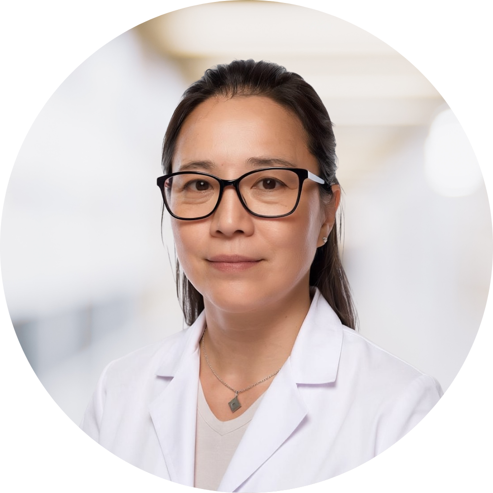 A woman wearing glasses and a white lab coat, standing indoors with a blurred background.