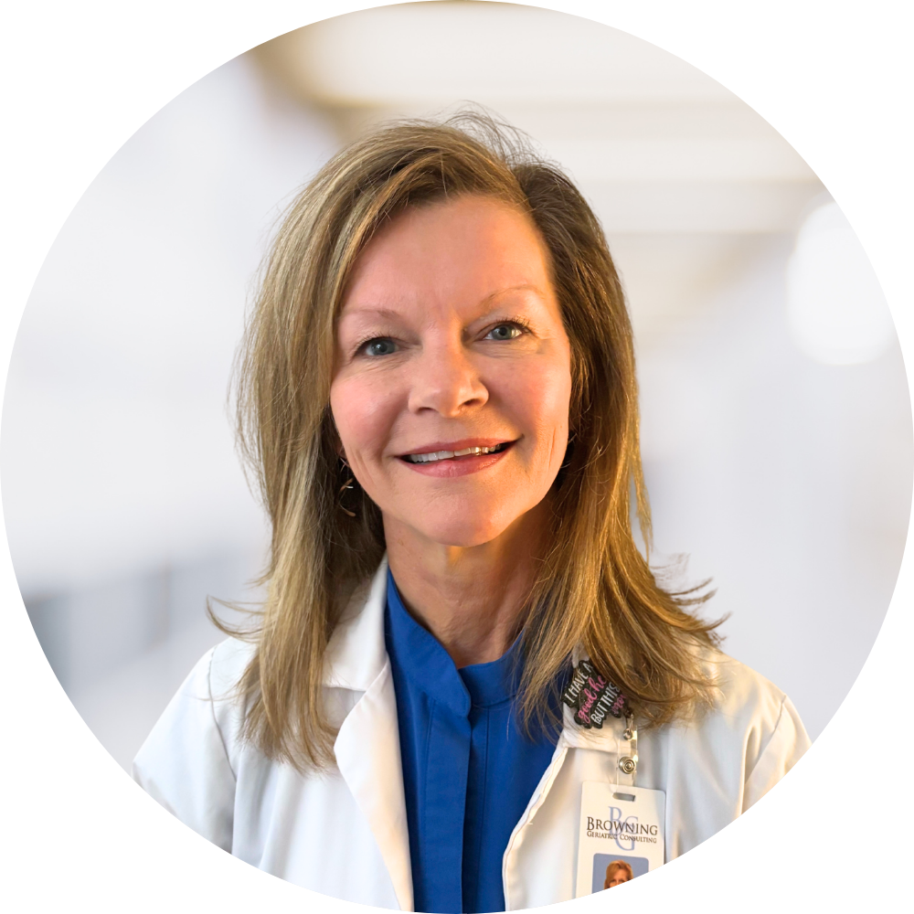 A smiling woman in a white medical coat with a blue shirt underneath, standing in a clinical or hospital environment.