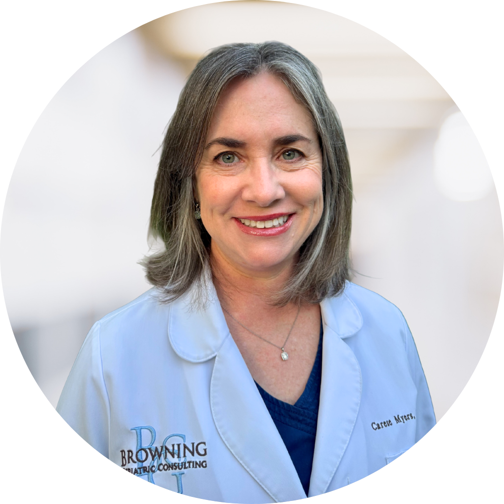 A smiling woman with shoulder-length gray hair wearing a white coat and a colorful floral blouse in a medical or professional setting.