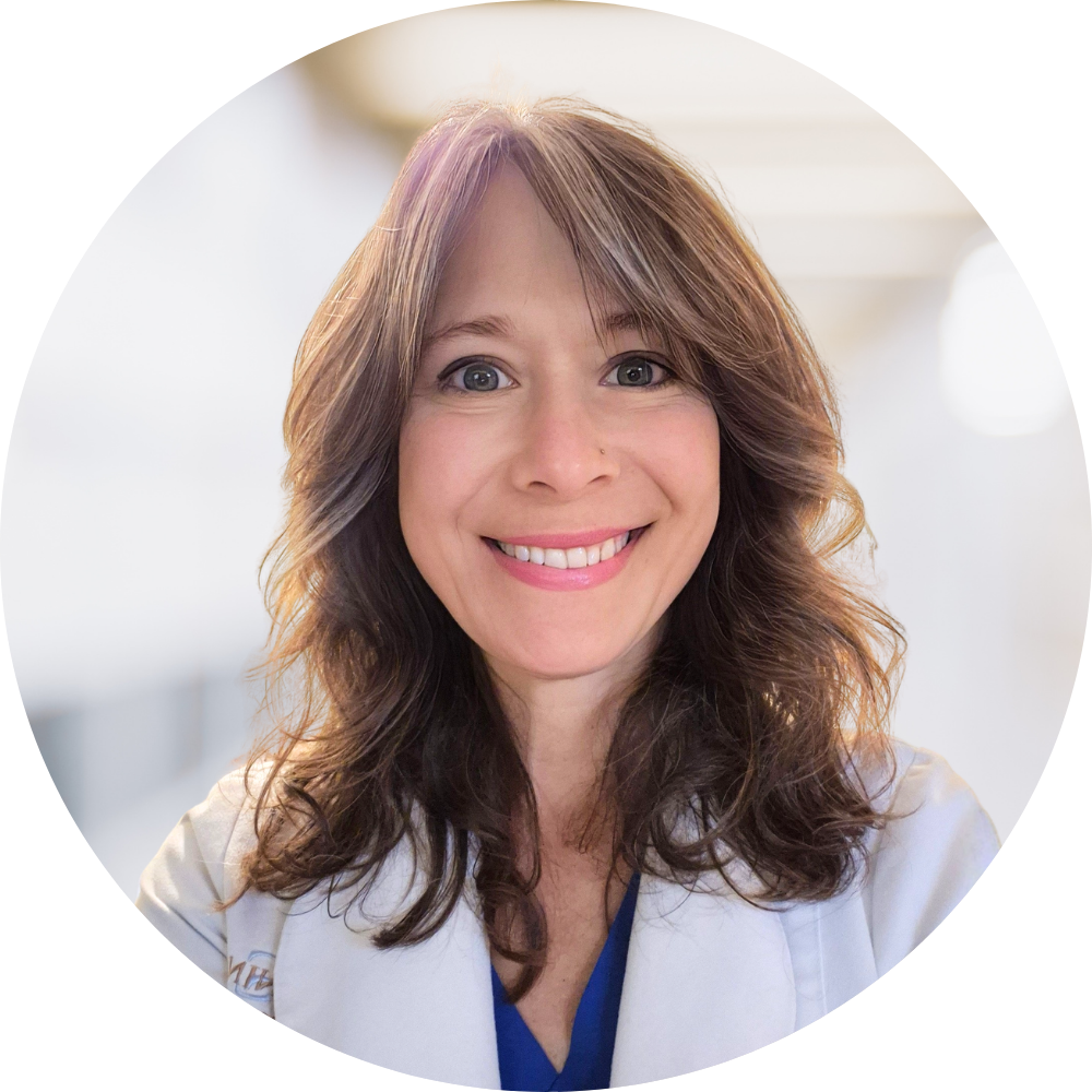 A smiling woman with wavy brown hair and a light complexion, wearing a white medical coat, in a professional setting.