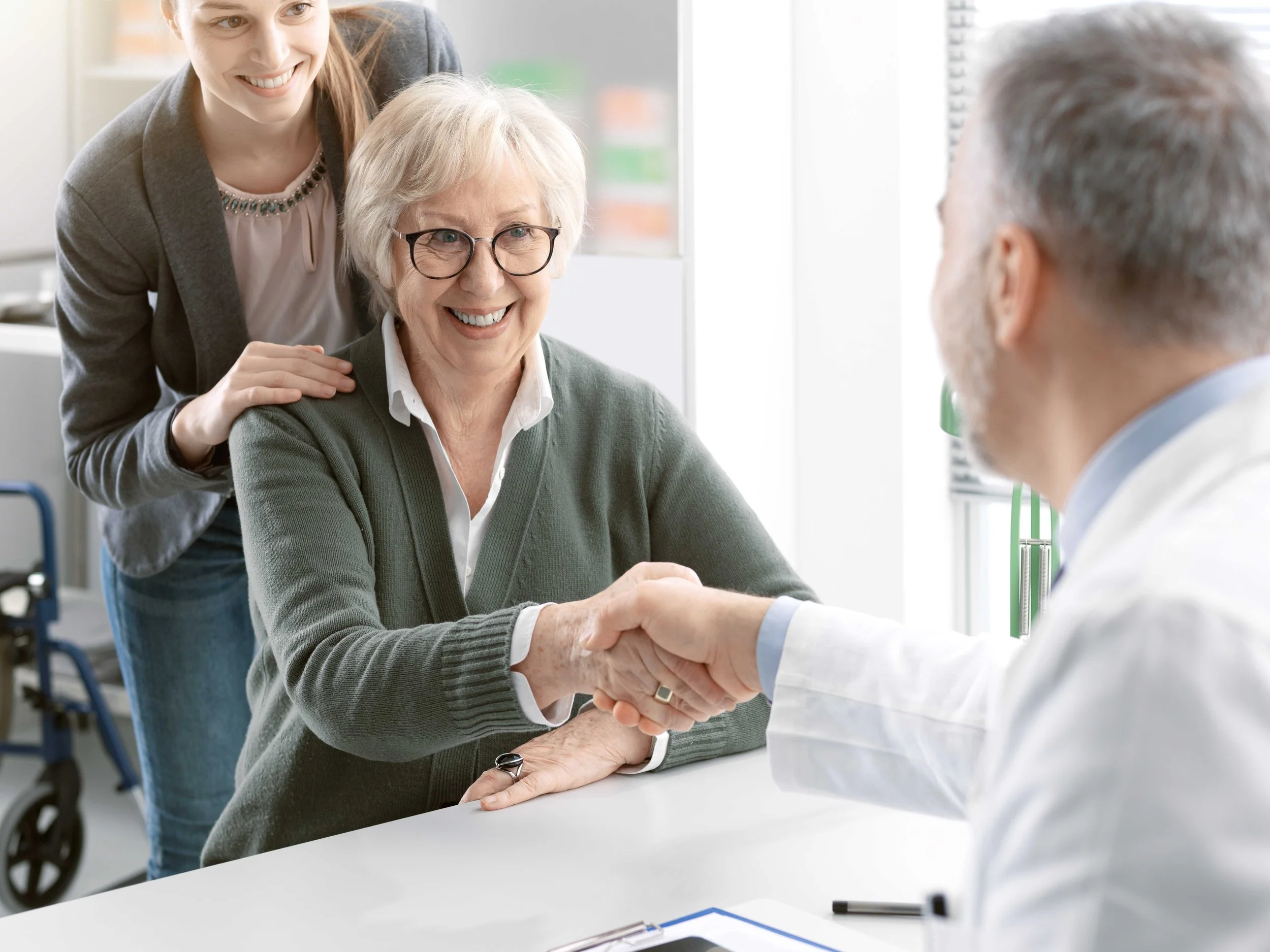 An elderly woman in glasses and a green sweater shaking hands with a doctor in a white coat inside a clinic or hospital, with a healthcare worker smiling and placing a hand on her shoulder.