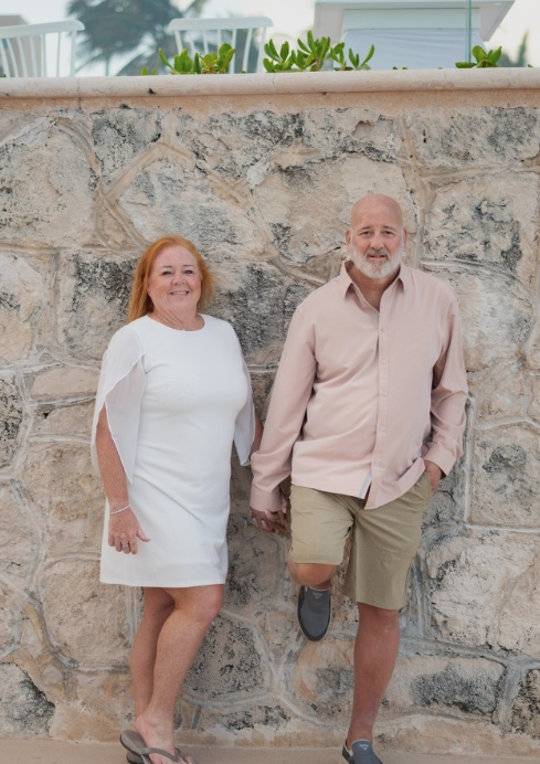 A smiling woman in a white dress and a man in a beige shirt and shorts holding hands, standing against a stone wall outdoors.