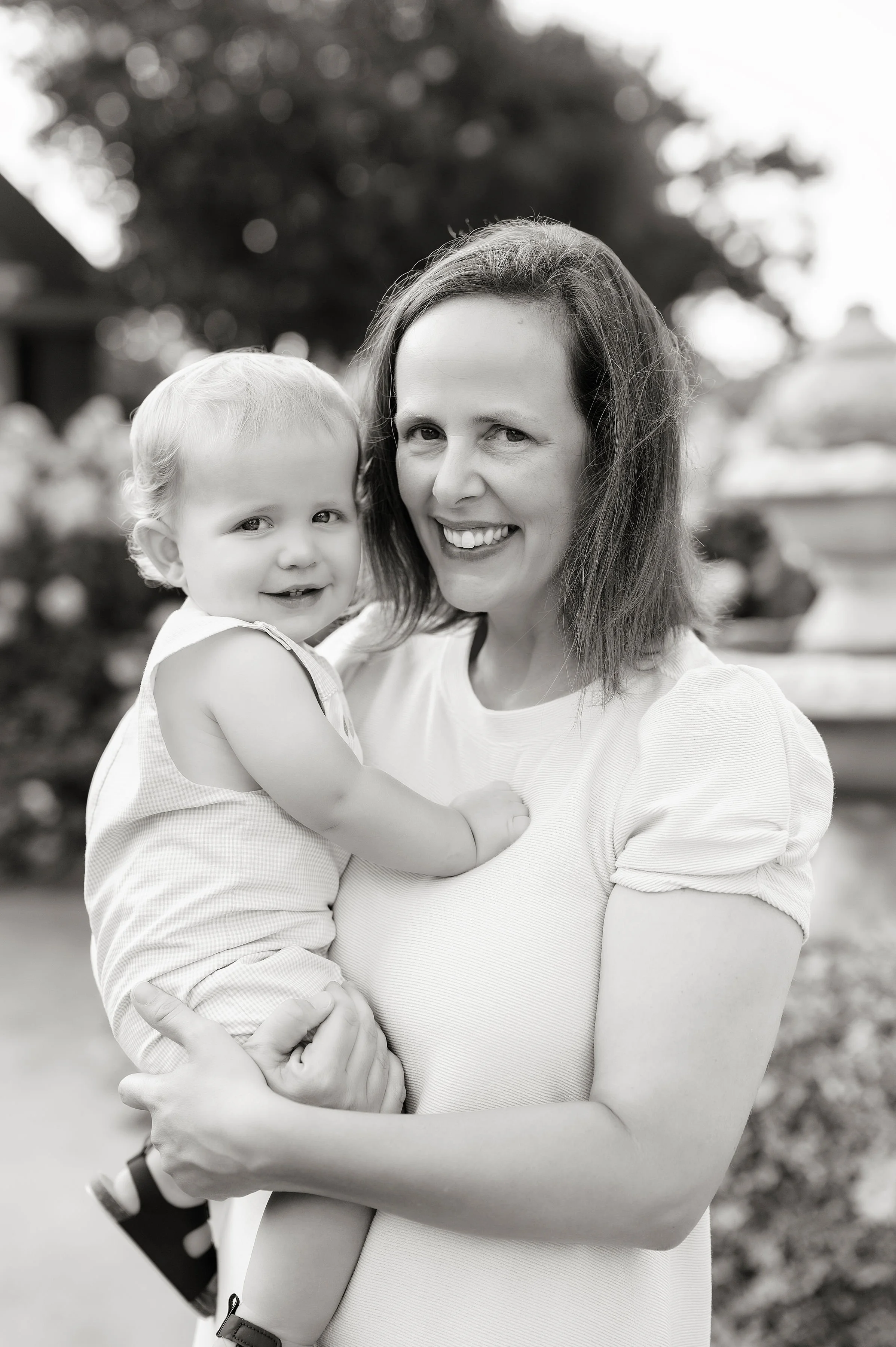 A smiling woman holding a young child in her arms outdoors, with trees and flowers in the background.