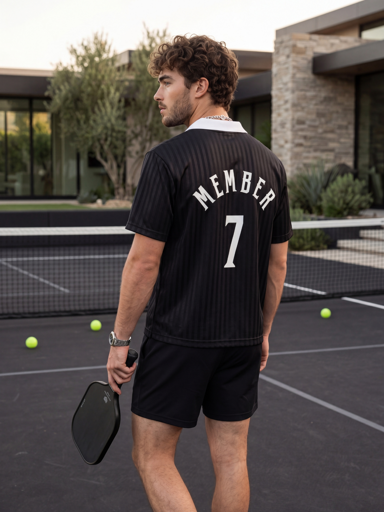 A young man on a tennis court holding a racket, wearing a black sports shirt with 'MEMBER' and the number 7 on the back, and black shorts. There are tennis balls on the court and a modern house in the background.