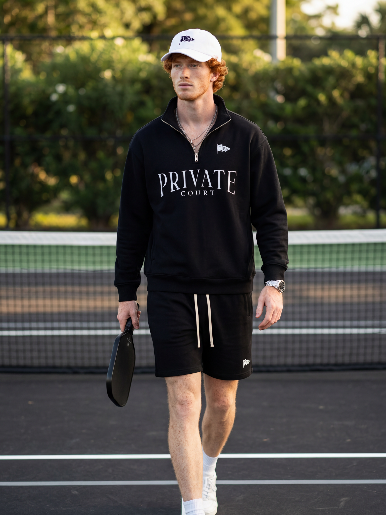 A young man with curly red hair and light skin walks on a tennis court. He wears a white cap with a logo, a black zip-up jacket with 'PRIVATE COURT' written on the front, black shorts, white socks, and sneakers. He is holding a paddle in his right ha