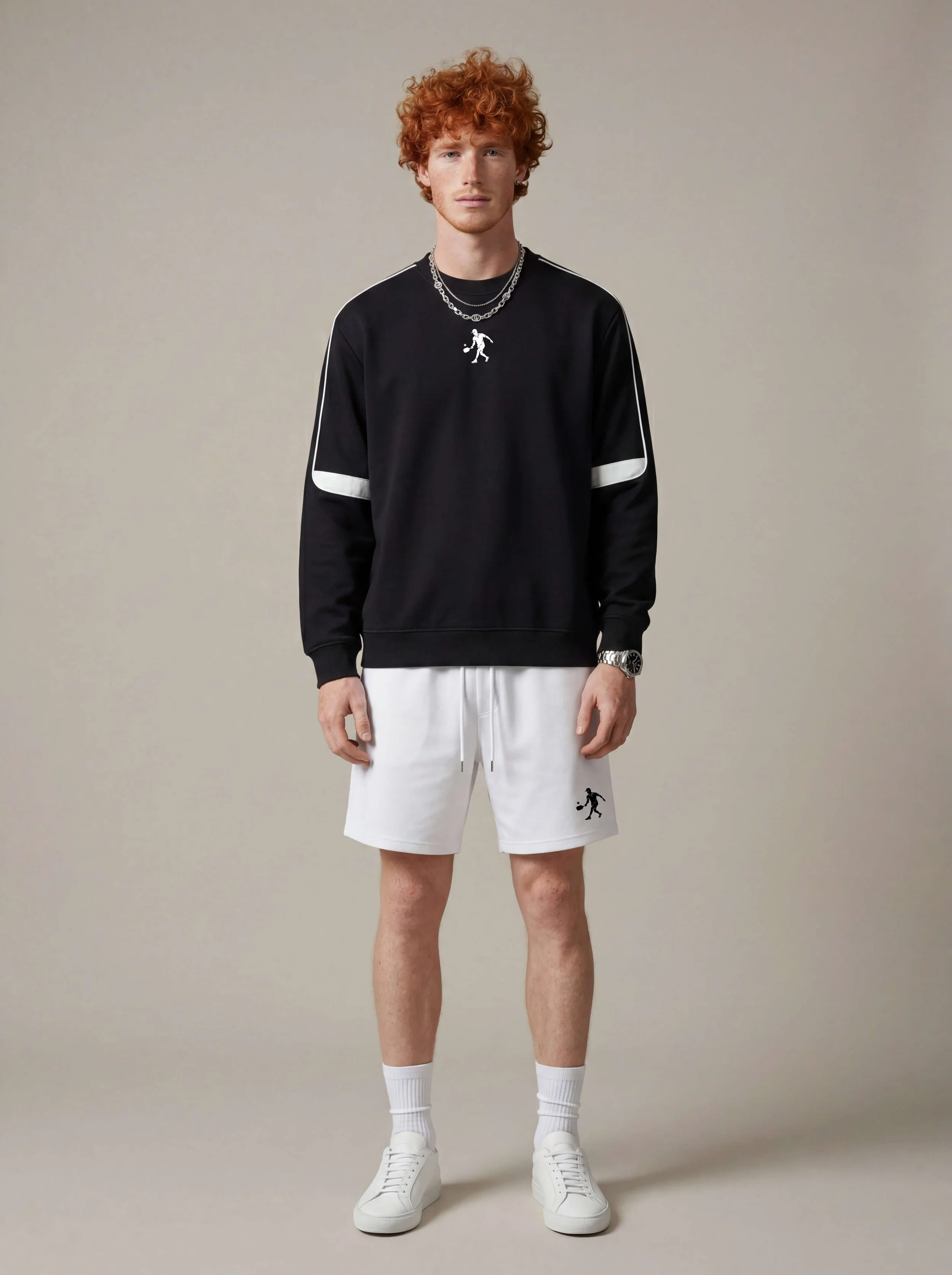 Male model wearing a luxury black pickleball crewneck sweatshirt with white athletic shorts and white sneakers in a minimalist editorial studio setting.