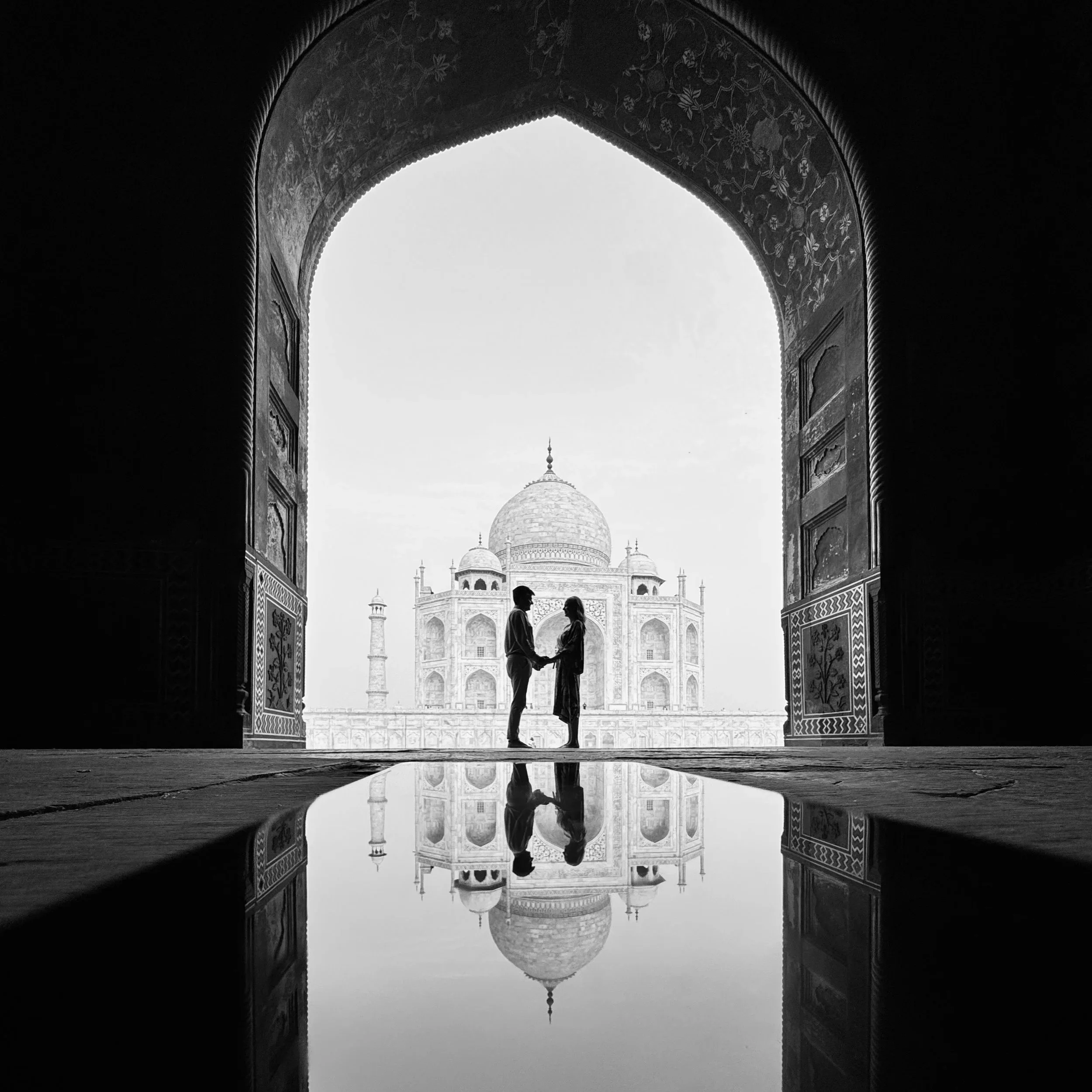 Silhouettes of a couple holding hands at the Taj Mahal with their reflection in a puddle, viewed from inside an ornate archway in black and white.