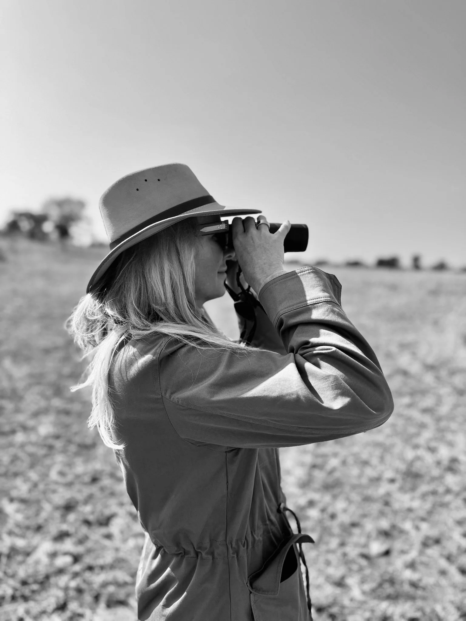A woman in a wide-brimmed hat and sunglasses is looking through binoculars in an open field.