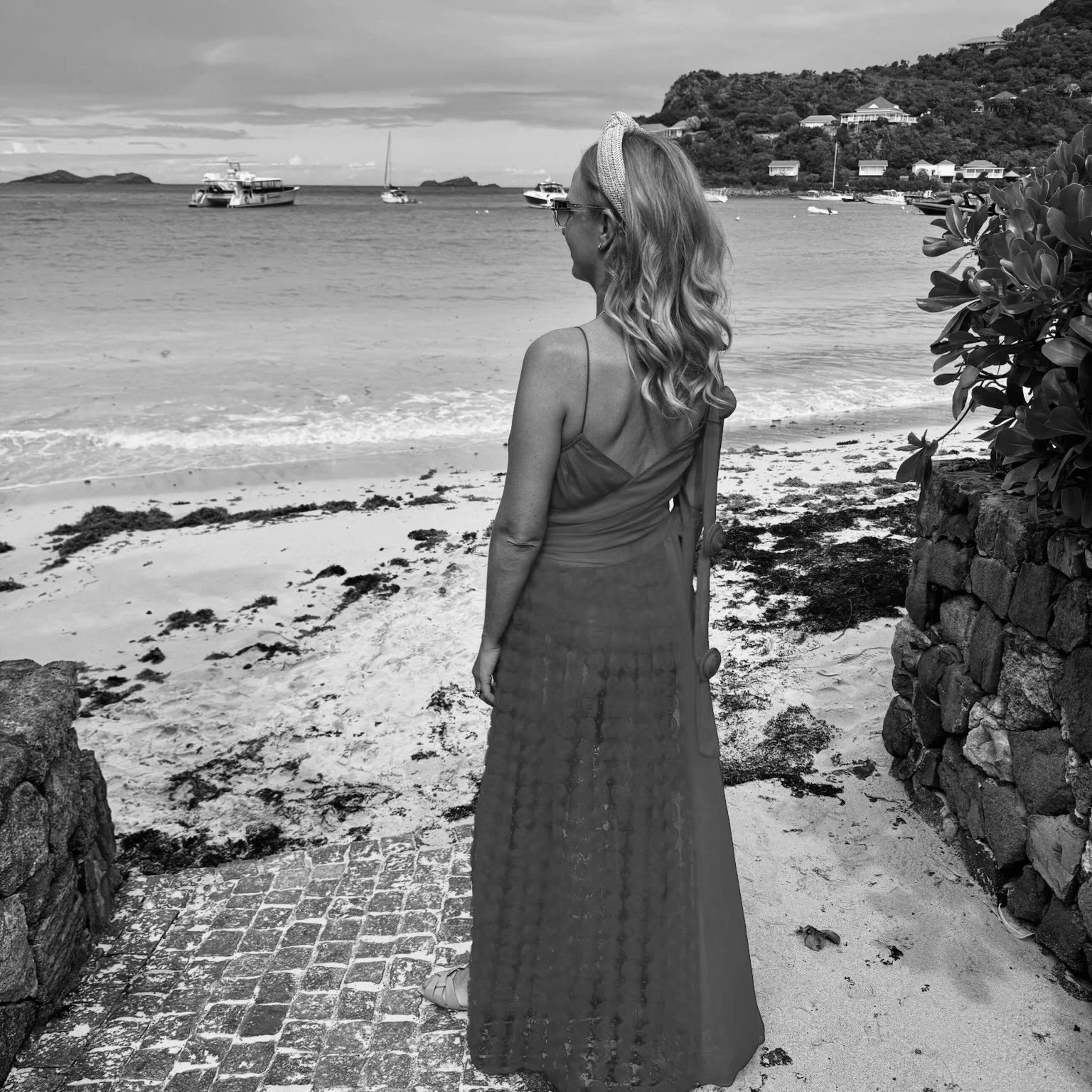 A woman with long wavy hair, wearing sunglasses, a headband, a sleeveless top, and a long skirt, standing on a beach near a stone wall, looking toward the water with boats and hills in the background.