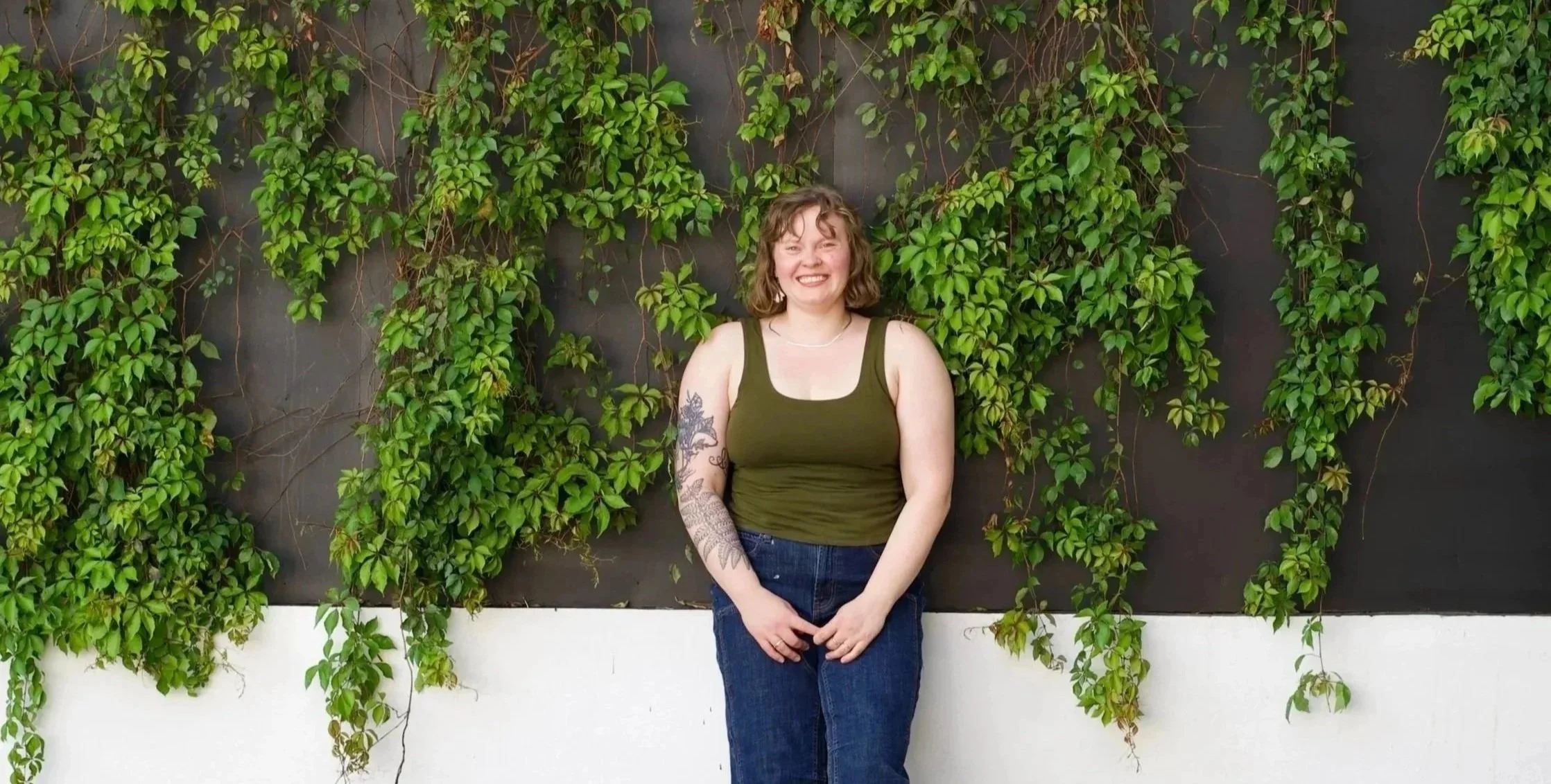 Olivia with curly hair and tattoos on her arm standing in front of a black wall with green ivy climbing on it, smiling at the camera.